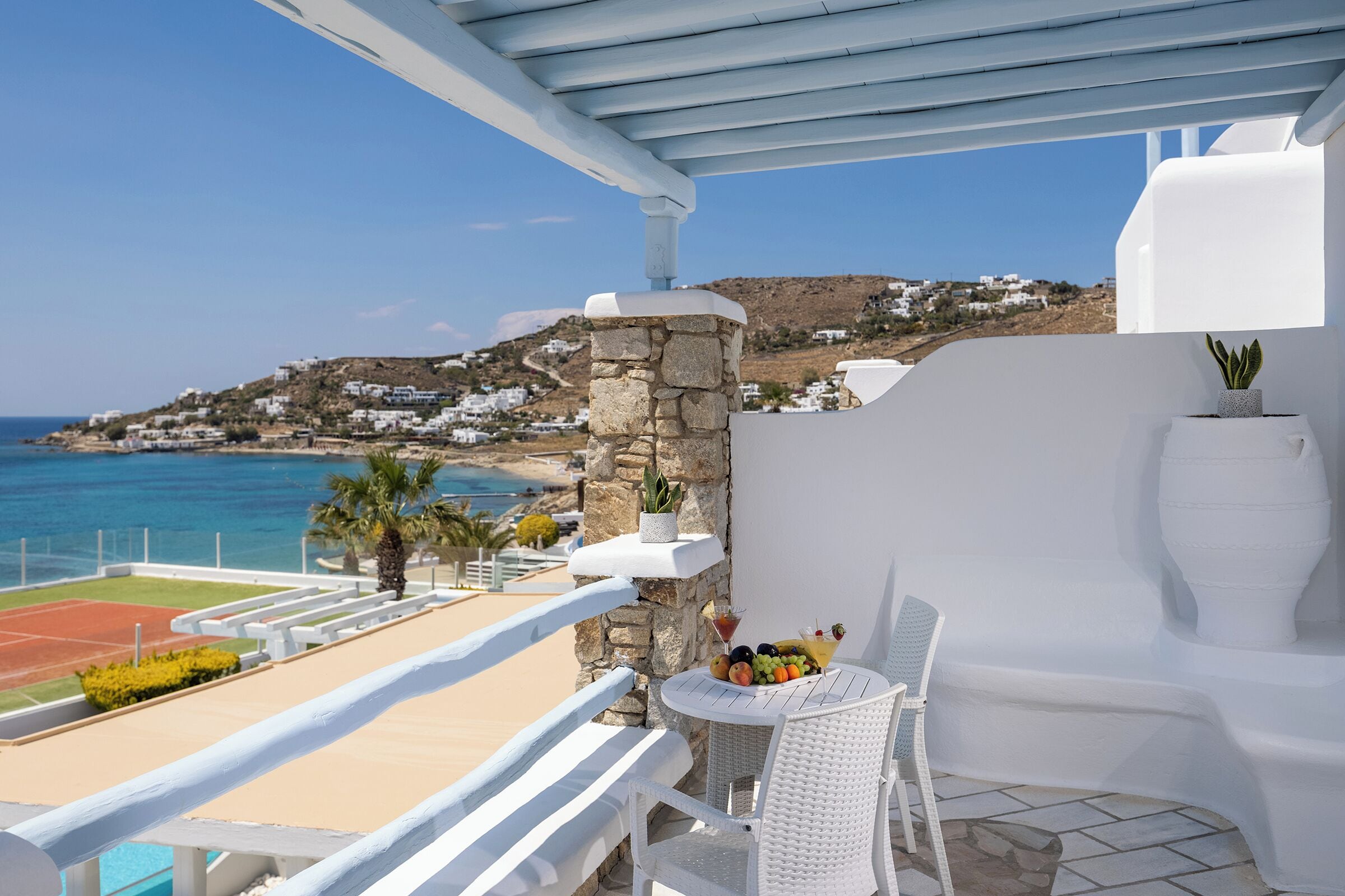 A balcony with a white wicker table and chair, a stone pillar, and a view of the sea and hillside houses.