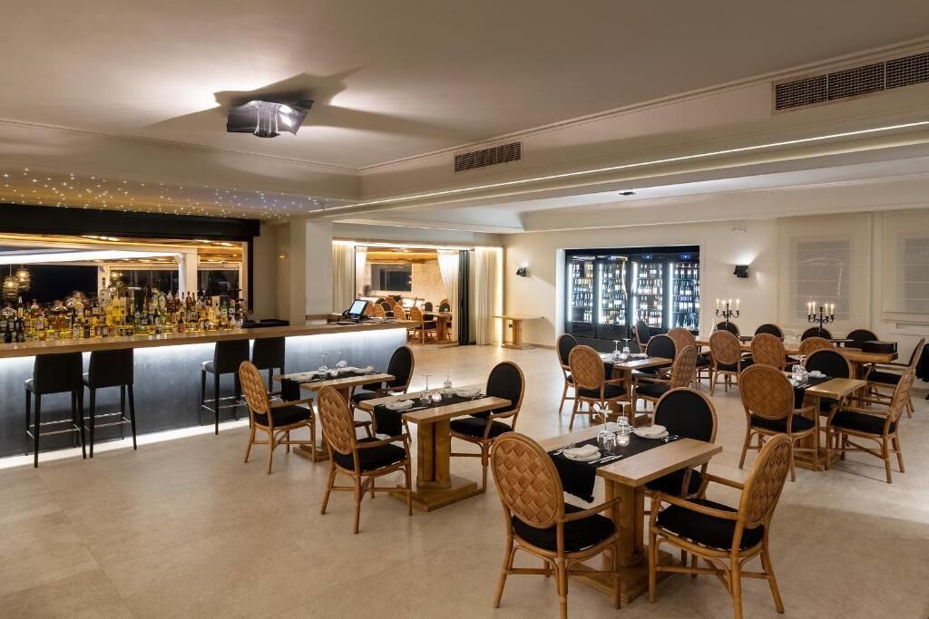 A spacious dining area with multiple tables and chairs, a bar with a variety of bottles, and a view of the city through large windows.
