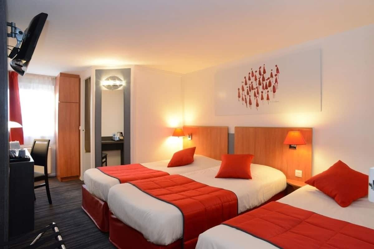 A hotel room with three beds, each with red bedding and white pillows. There is a desk with a chair, a TV mounted on the wall, and a large mirror. The room has a modern design with a warm color scheme.