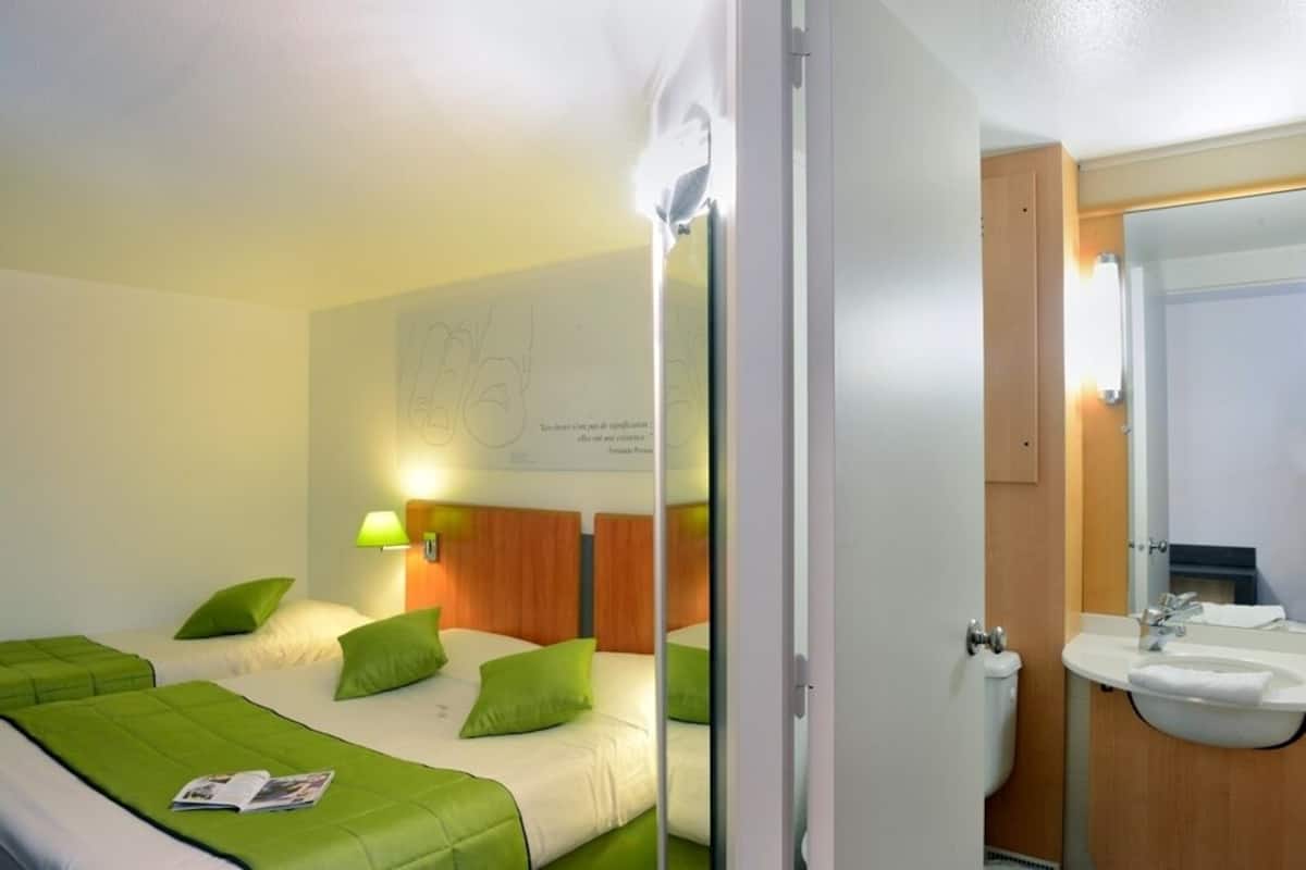 A hotel room with two beds, a wooden headboard, a bedside lamp, a bathroom with a sink and mirror, and a door leading to another room.