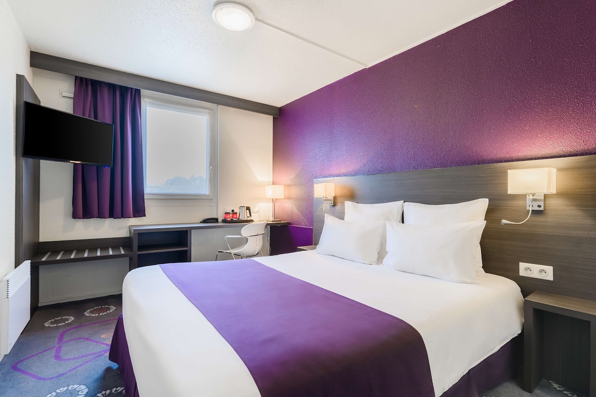 A hotel room with a bed, a desk, a chair, a TV, and a window with curtains.