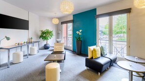Een moderne hotelkamer met een bank, poufs, een kleine tafel en een bureau met een lamp. Er is een groot raam met uitzicht op buiten.
