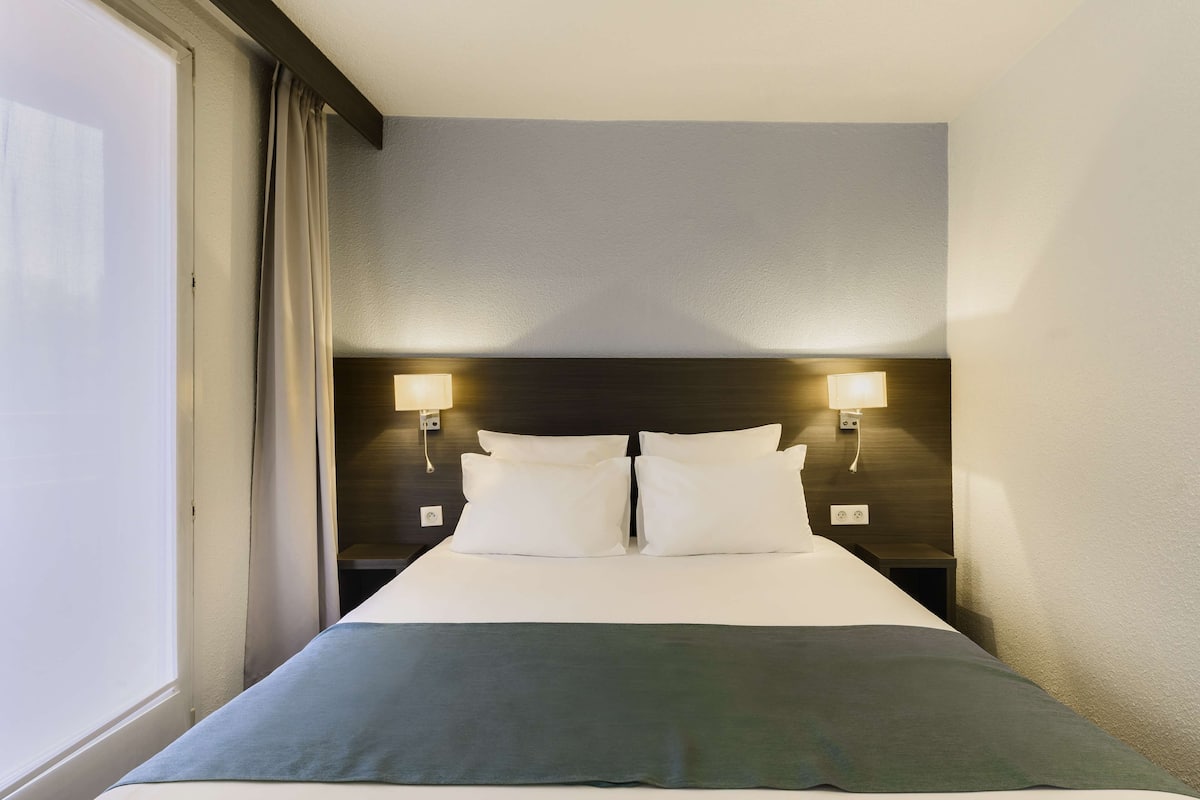 A hotel room with a large bed, two bedside lamps, and a window with curtains.