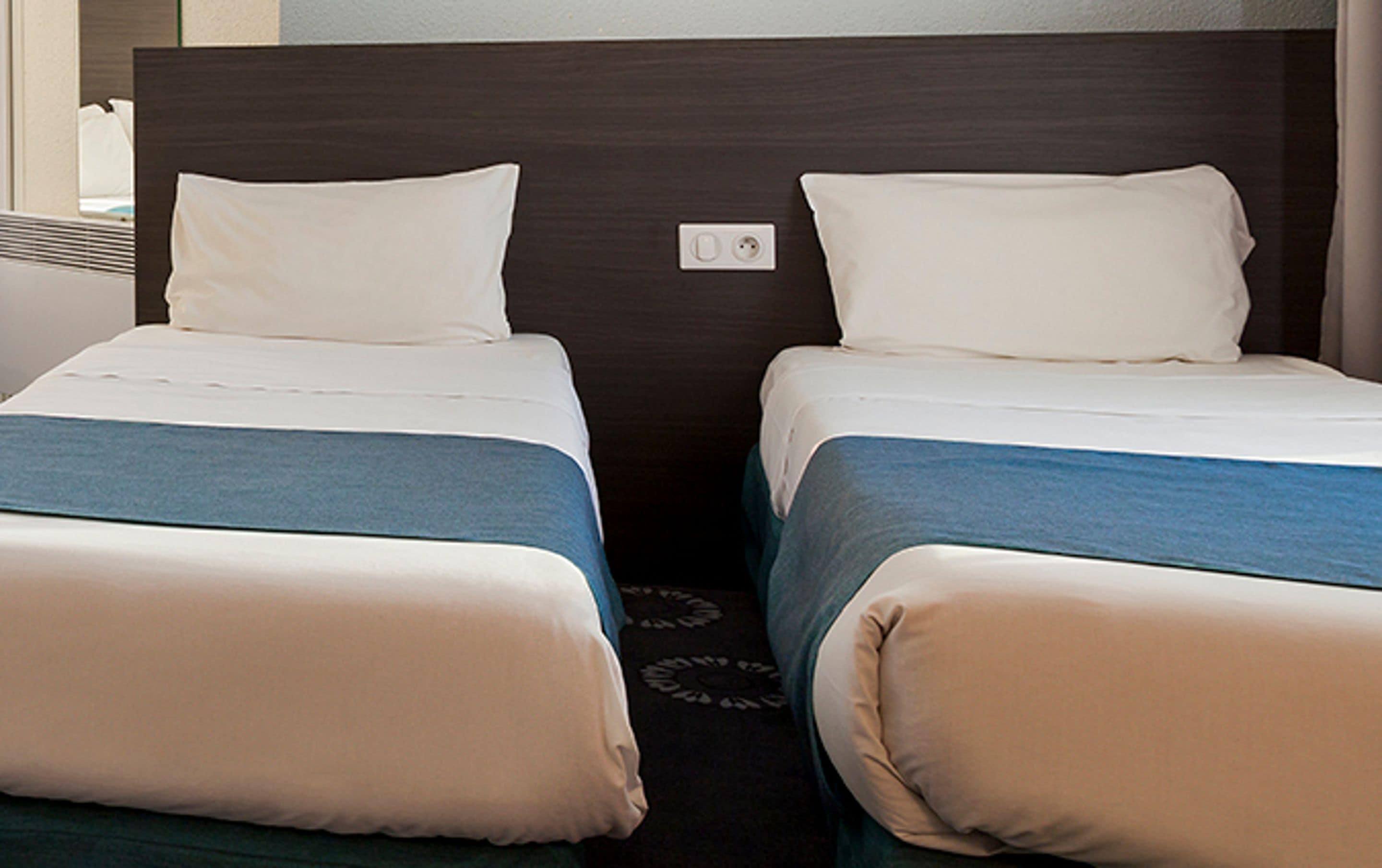 A hotel room with two single beds, each with a white pillow and a blue bedspread.