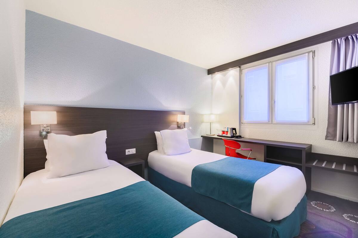 A hotel room with two beds, a desk, a chair, a TV, and a window with curtains.