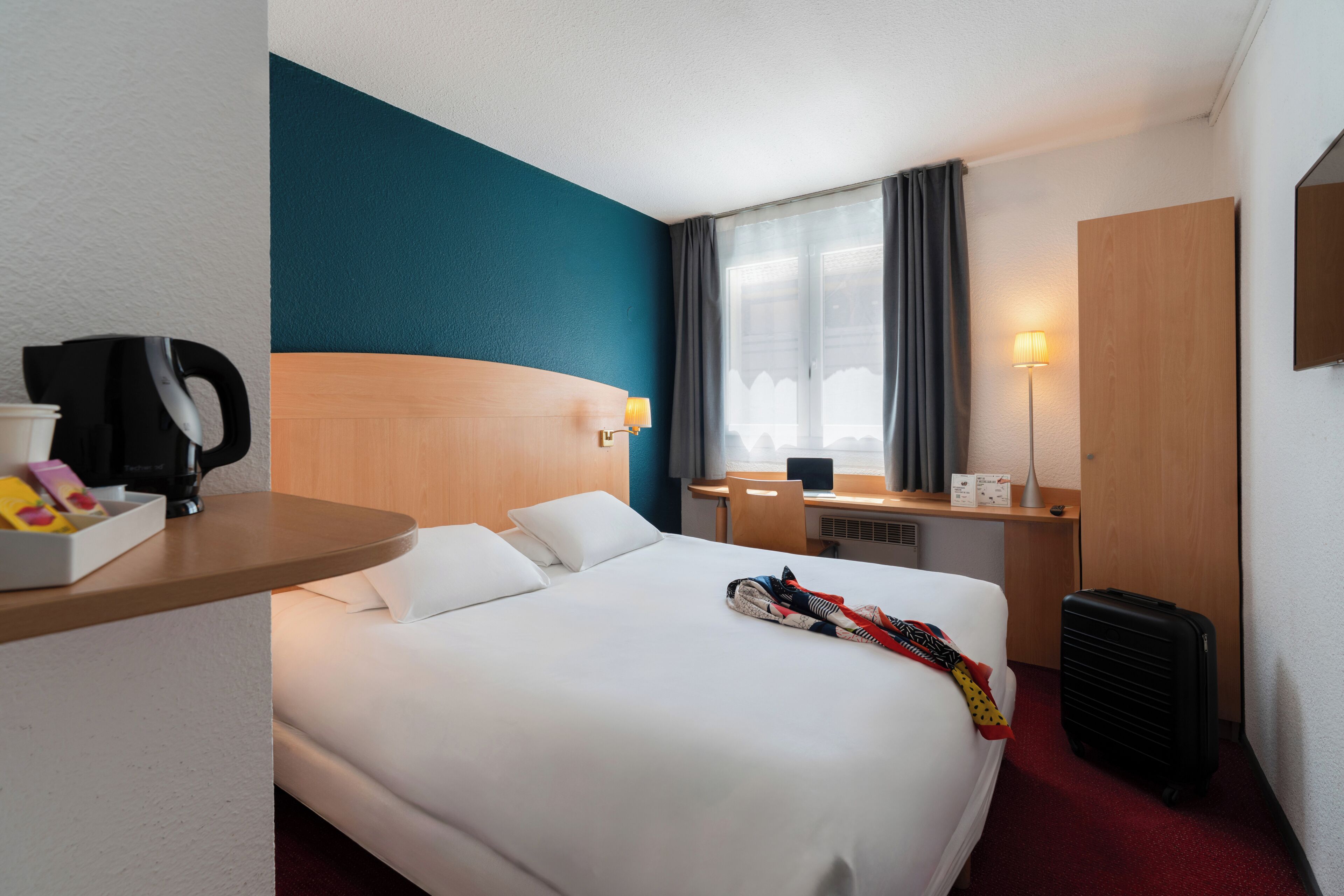 Una habitación de hotel con cama, escritorio, silla, cafetera, lámpara y maleta.
