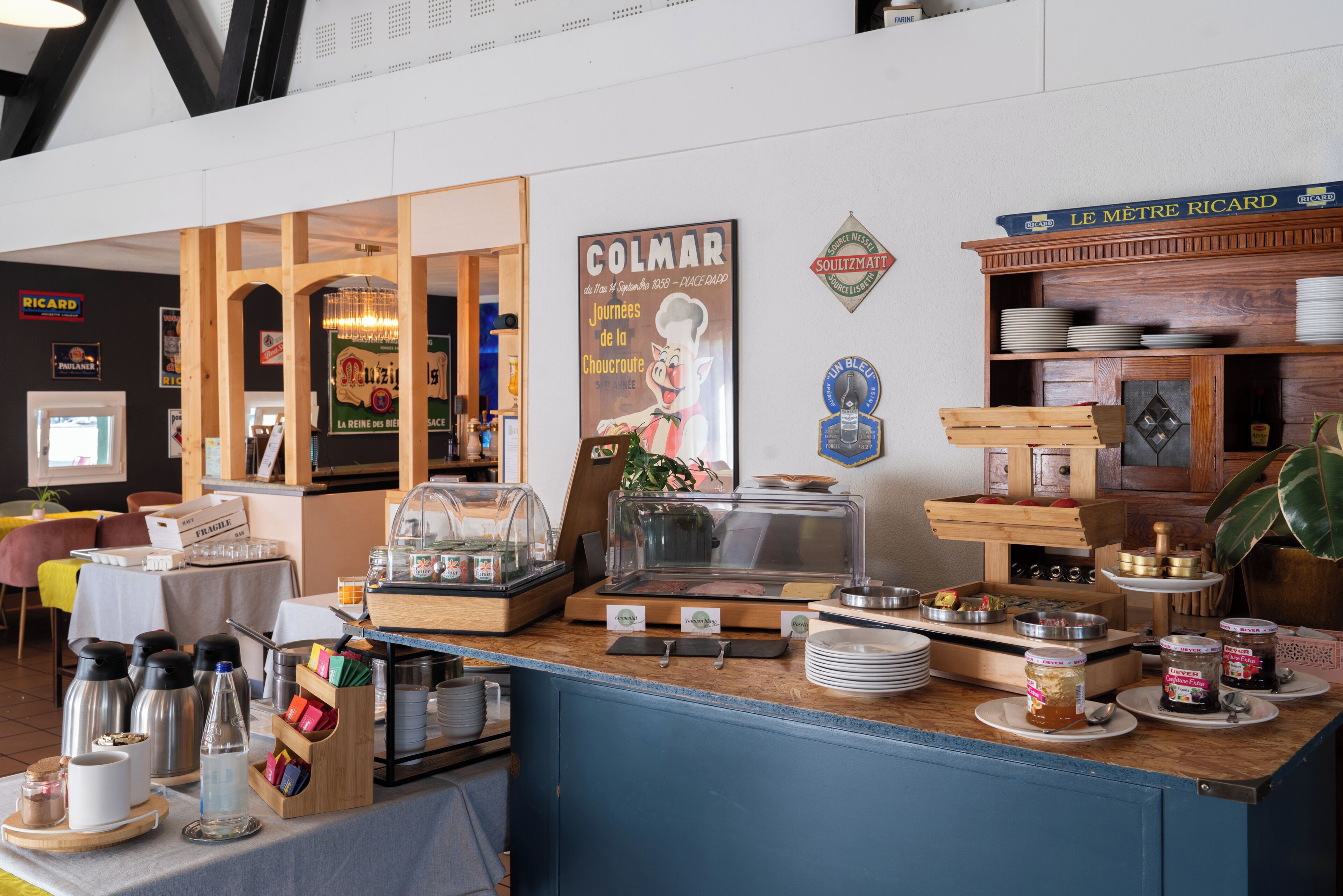 A breakfast bar with a variety of food items and condiments, a poster of a chef, and a sign for 'Le Maitre Ricard' wine.
