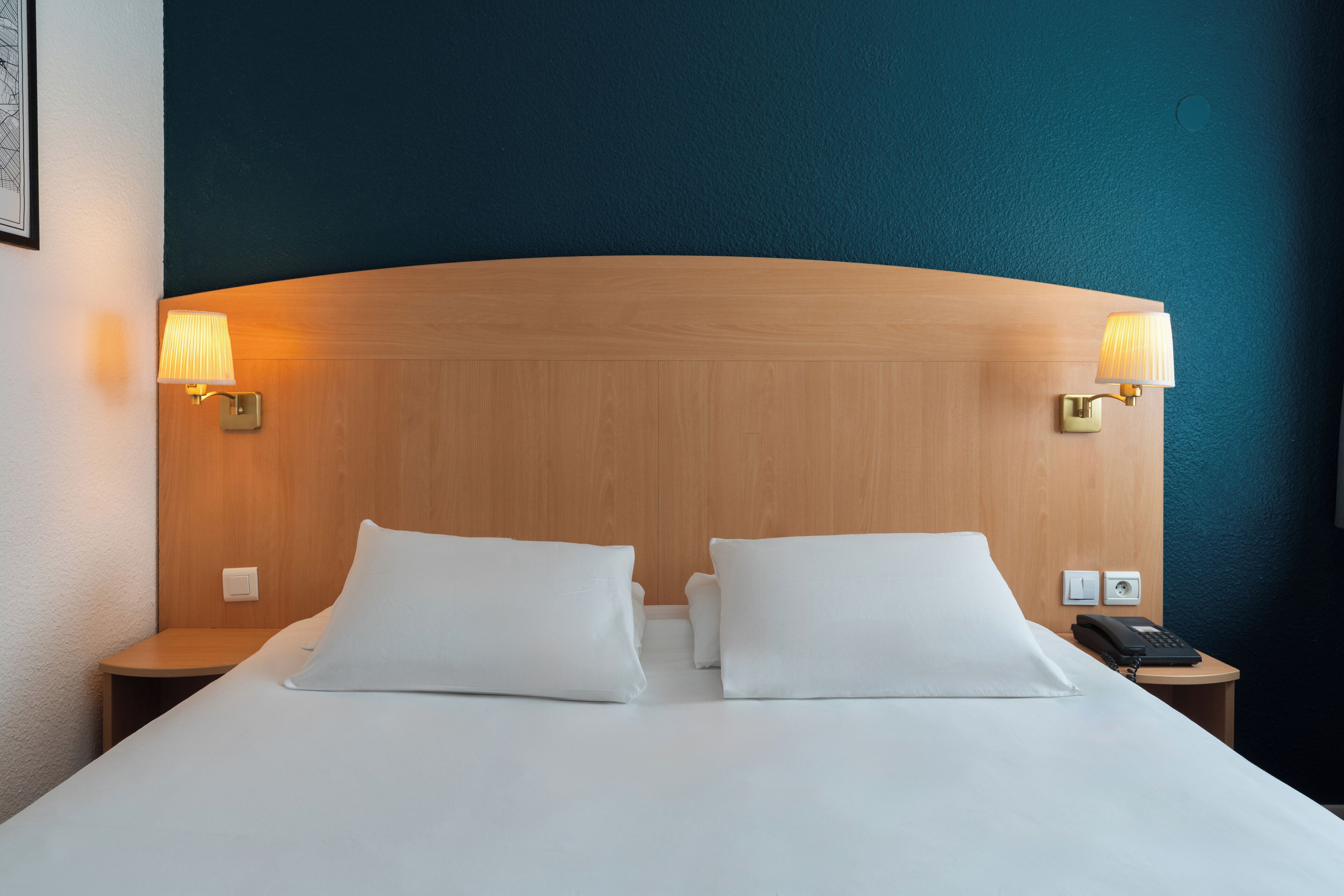 Une chambre d’hôtel avec une tête de lit en bois, deux lampes de chevet, un lit avec du linge de maison blanc et une table de chevet avec un téléphone.
