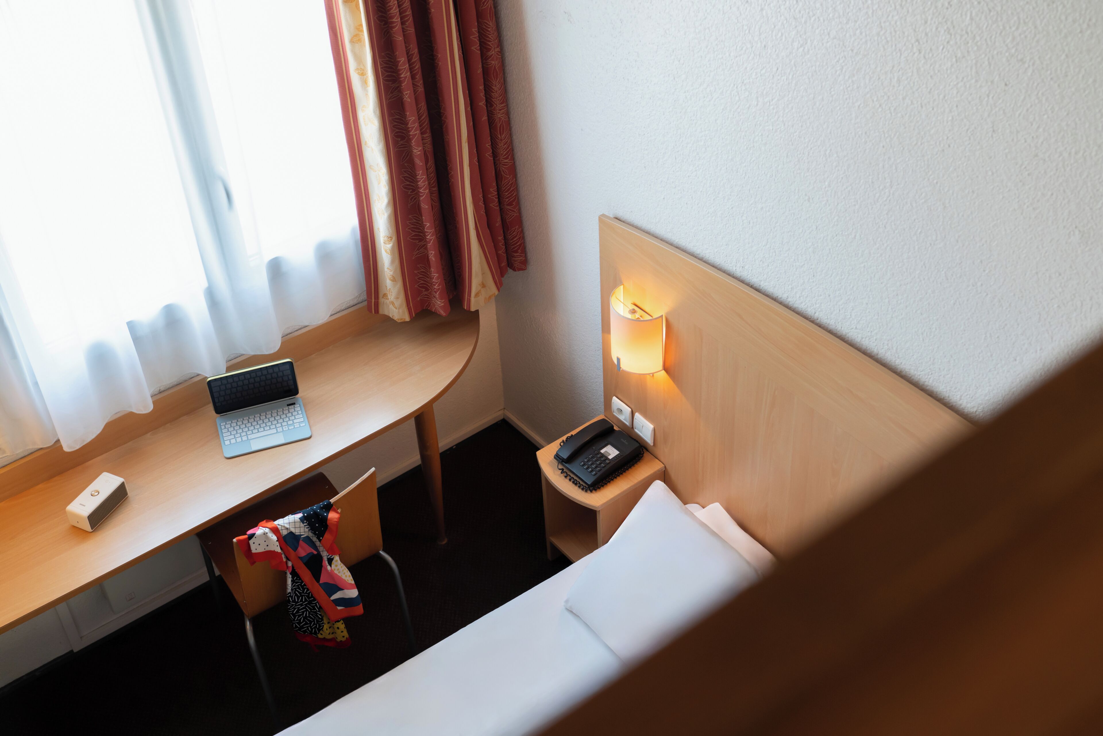 A hotel room with a desk, laptop, phone, and a bedside lamp.