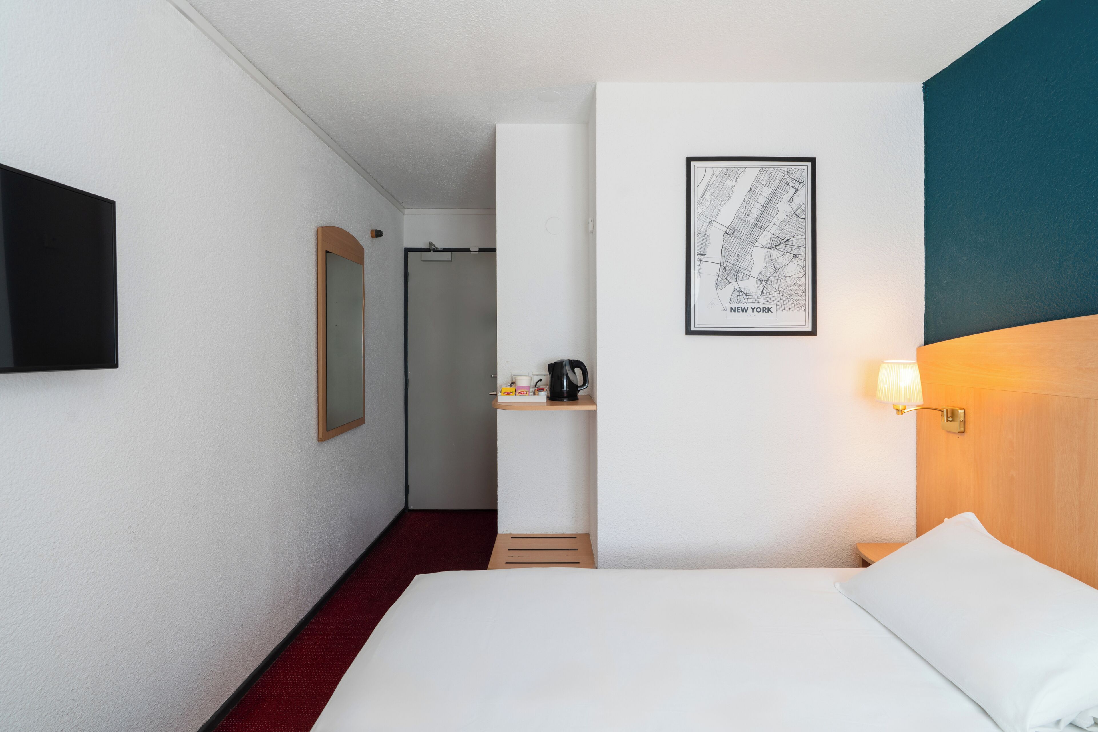 A hotel room with a bed, a wall-mounted TV, a framed map, and a nightstand with a coffee maker.