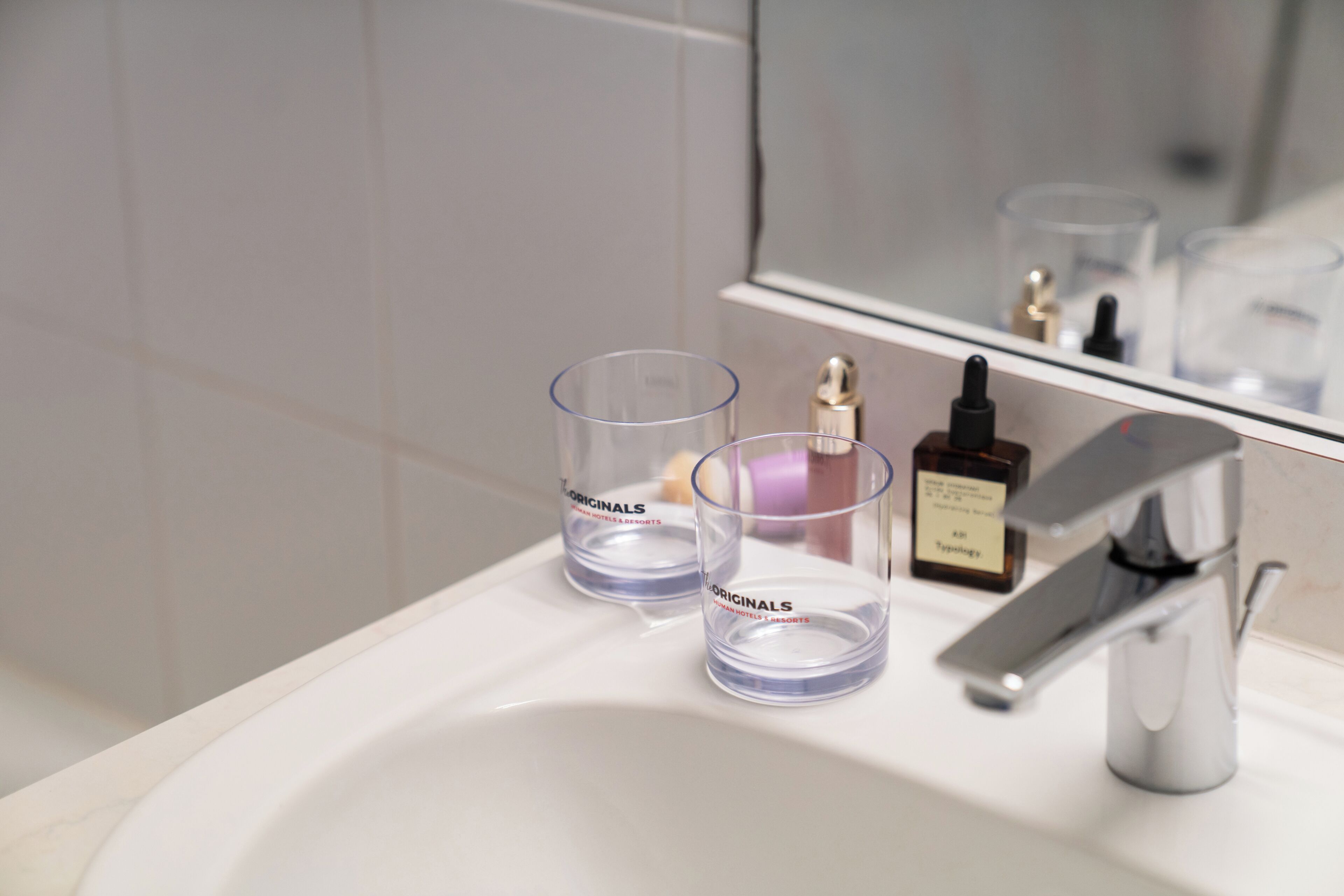 Un lavamanos de baño con dos vasos de vidrio transparentes etiquetados 'The Originals' y una botella de perfume.
