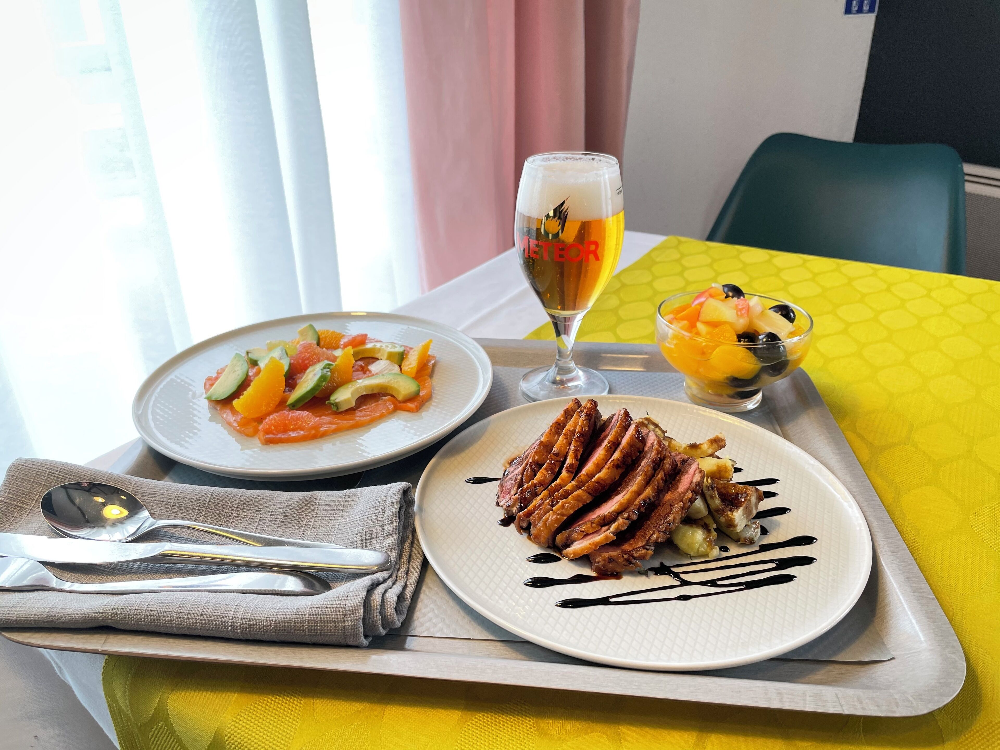 A tray with a plate of steak, a salad, and a fruit bowl, a glass of beer, and cutlery on a yellow tablecloth.