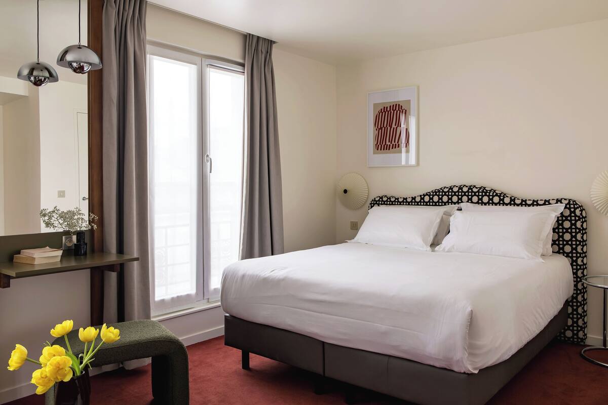 A neatly arranged bedroom with a bed, a bedside table, a chair, a window with curtains, and a wall-mounted artwork.