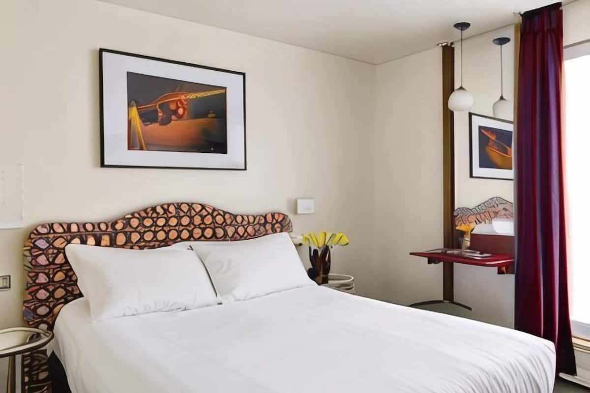 A neatly made bed with white linens, a framed picture above it, and a bedside table with a lamp and vase of flowers.