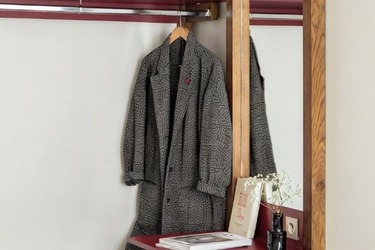 Una chaqueta colgada en una percha de madera, una mesita con un libro y un jarrón de flores secas, y un espejo con marco de madera.