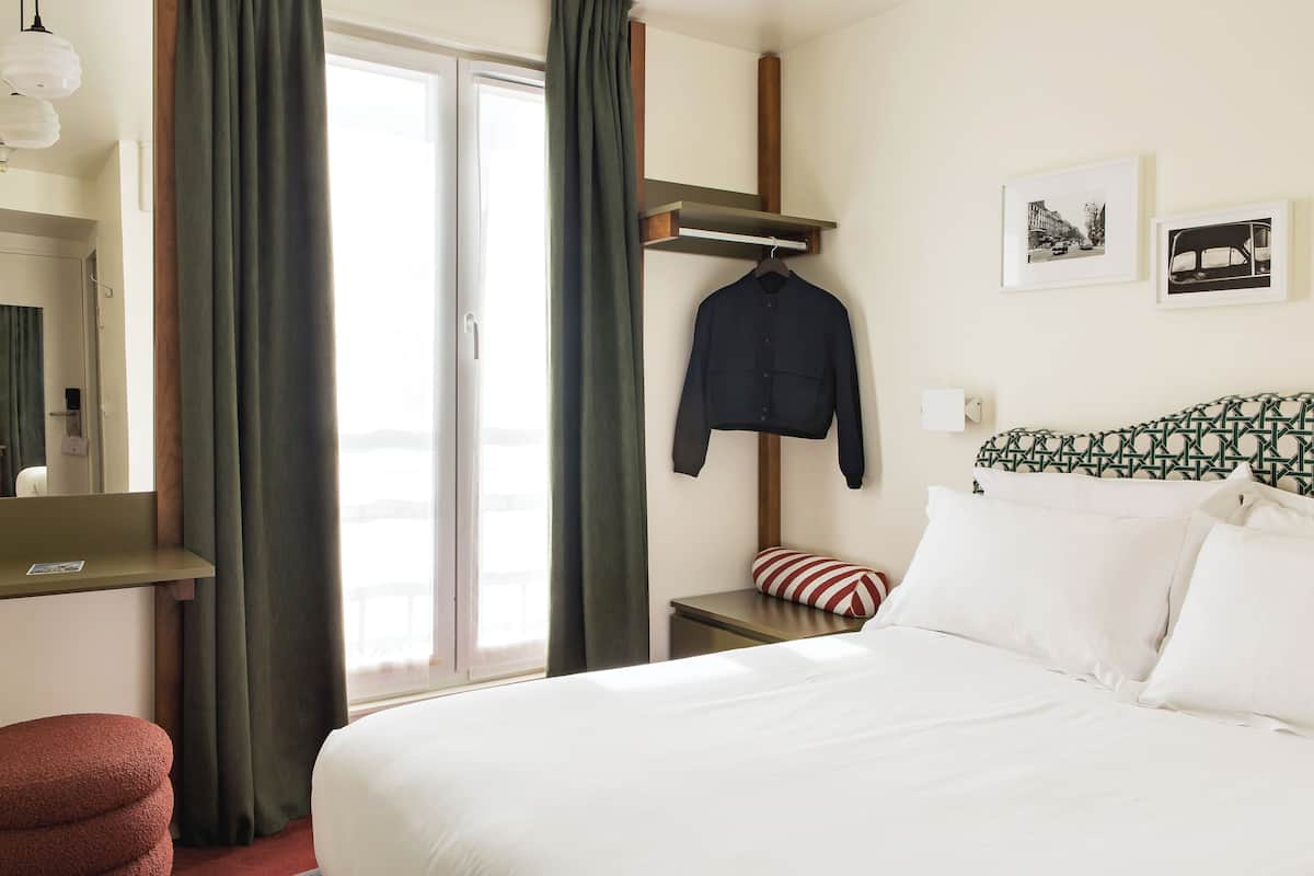 A hotel room with a bed, a bedside table, a hanging coat, and a window with curtains.