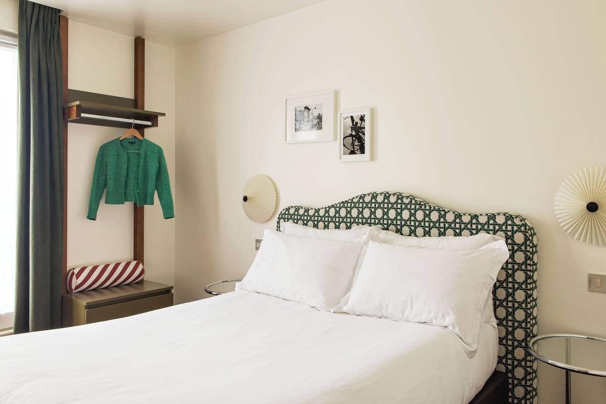 A neatly made bed with white linens, a patterned headboard, and a bedside table with a red and white striped pillow.