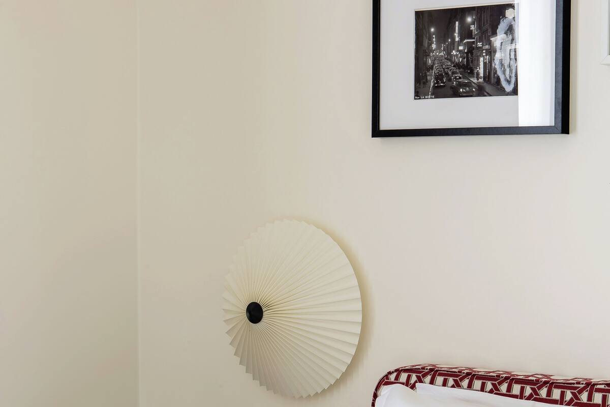 A neatly made bed with white linens, a bedside table with a vase of yellow flowers, a wall-mounted fan, and a framed picture on the wall.