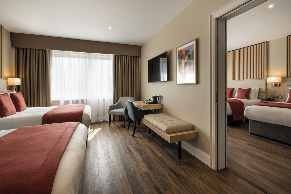 A hotel room with two beds, a desk, a chair, a bench, and a television.