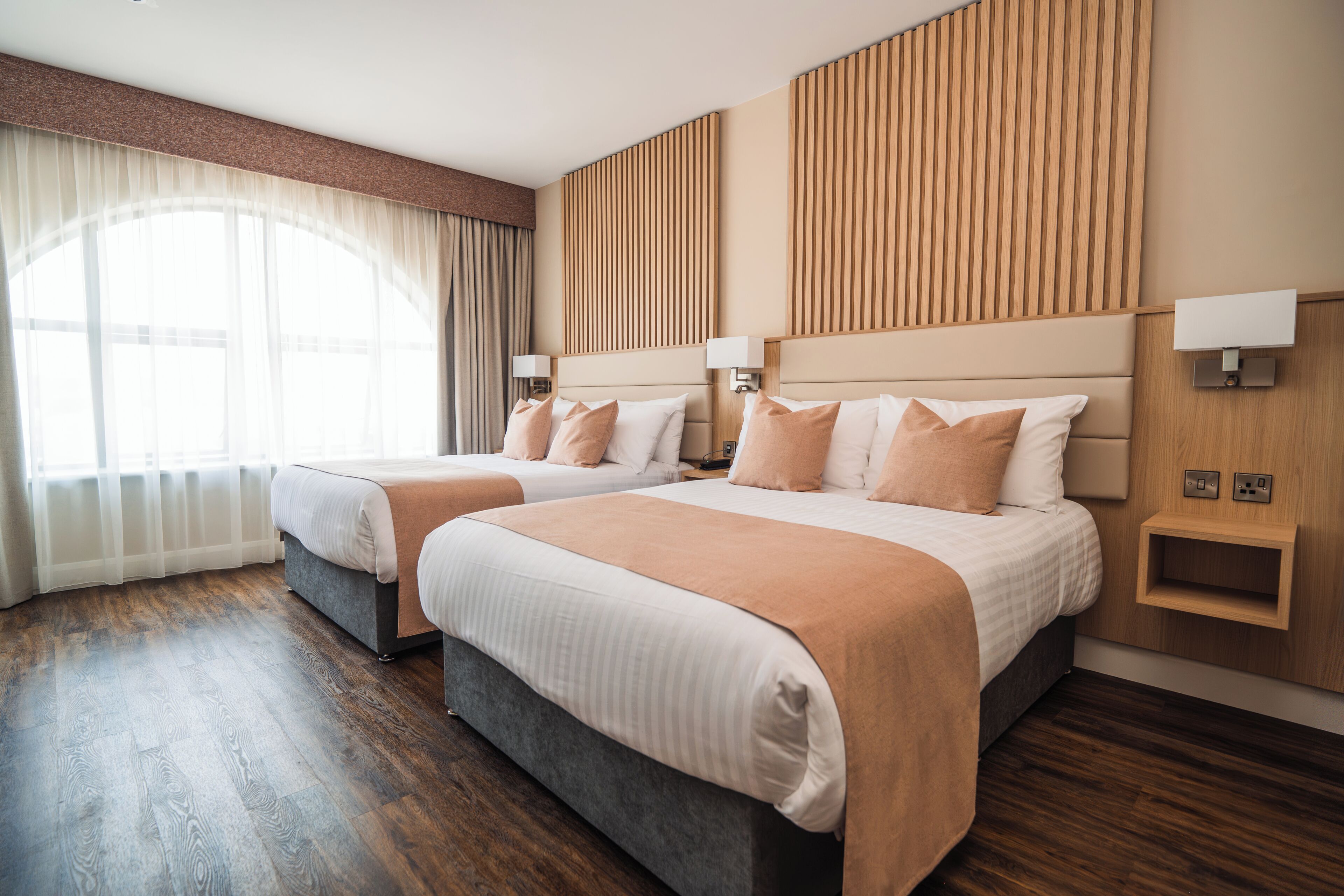 A hotel room with two beds, wooden paneling on the walls, and a large window with sheer curtains.