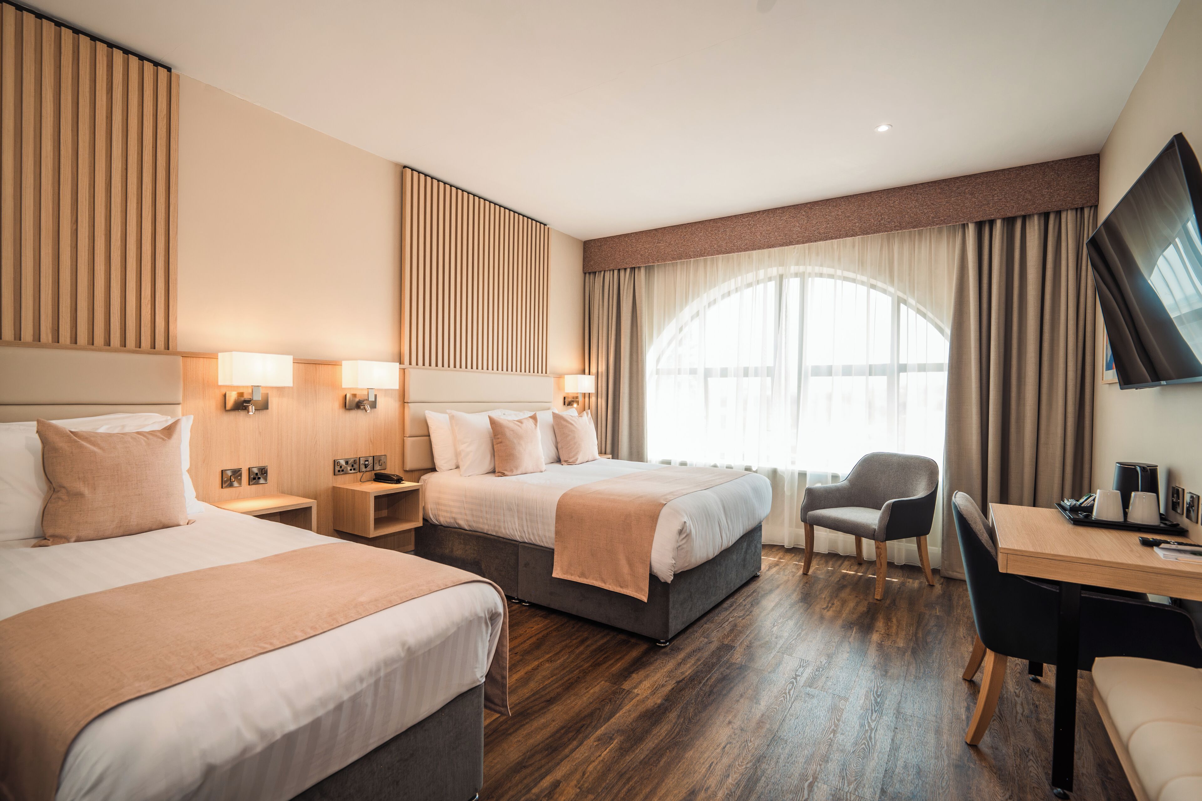 A hotel room with two beds, a desk, a chair, a TV, and a large window with curtains.
