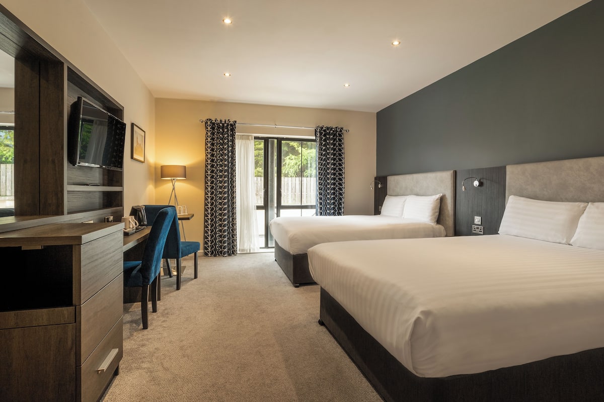 A hotel room with two beds, a desk with a chair, a TV, and a large window with curtains.