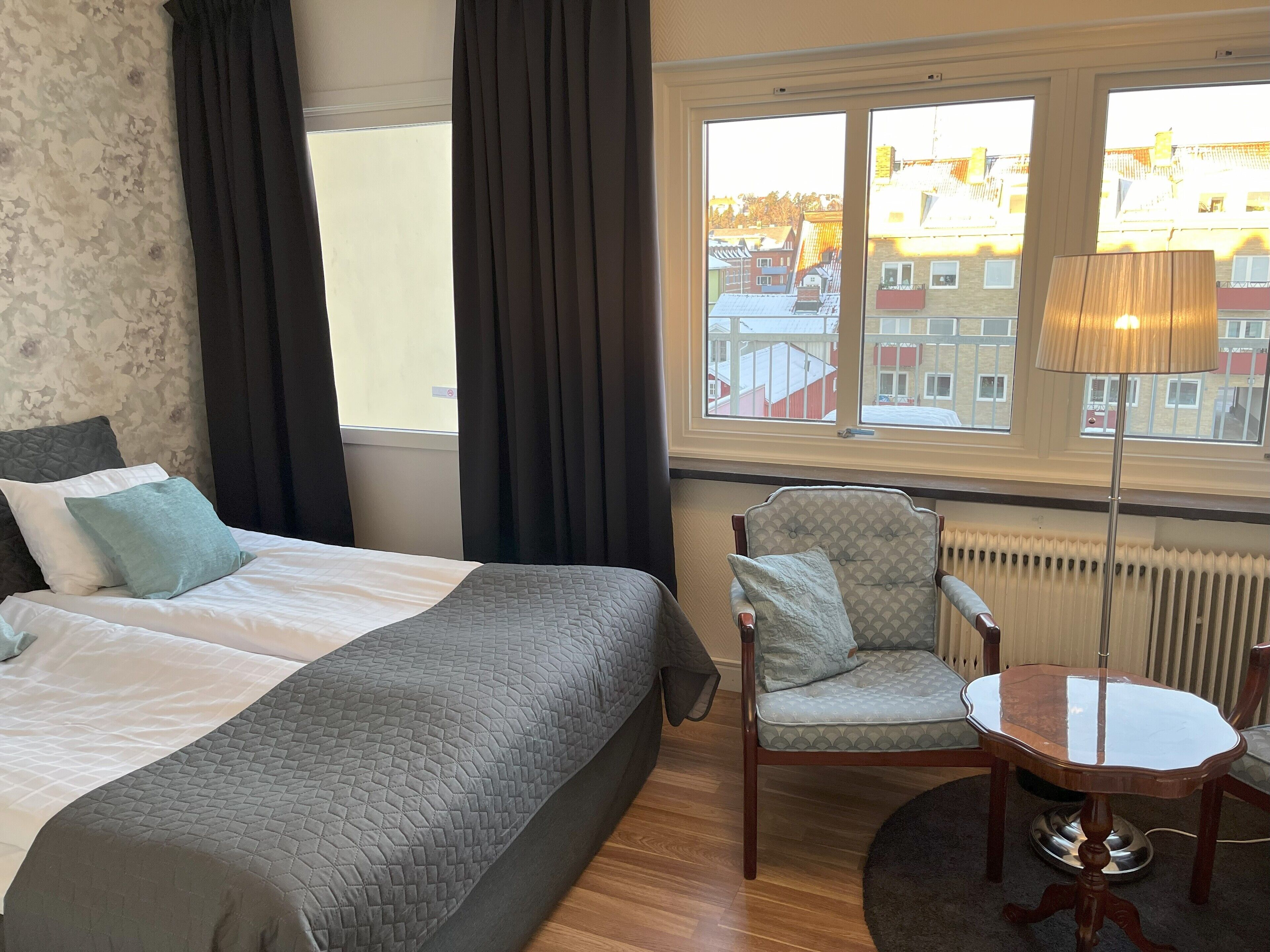 Ett hotellrum med en säng, en stol, ett bord och utsikt över byggnader genom fönstret.