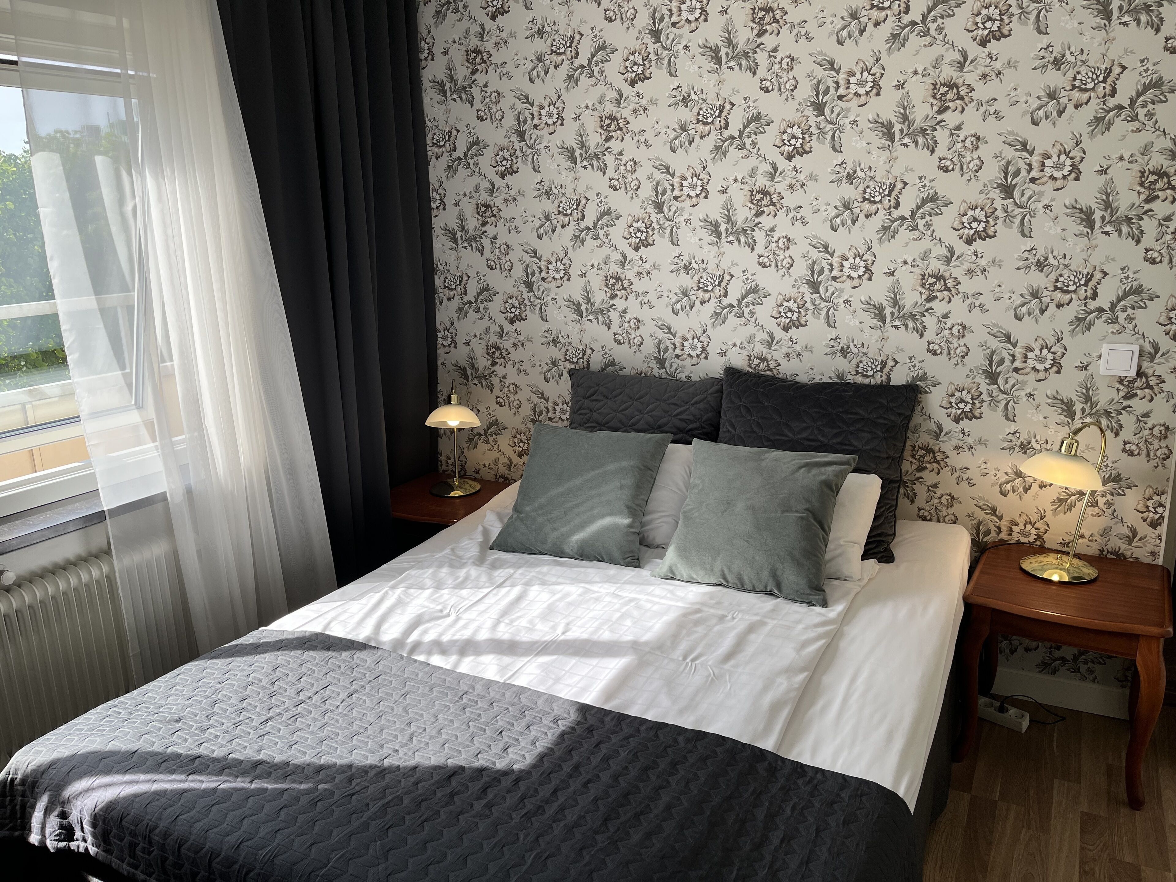 Una cama bien hecha con una colcha estampada, dos lámparas de noche y papel tapiz con motivos florales.