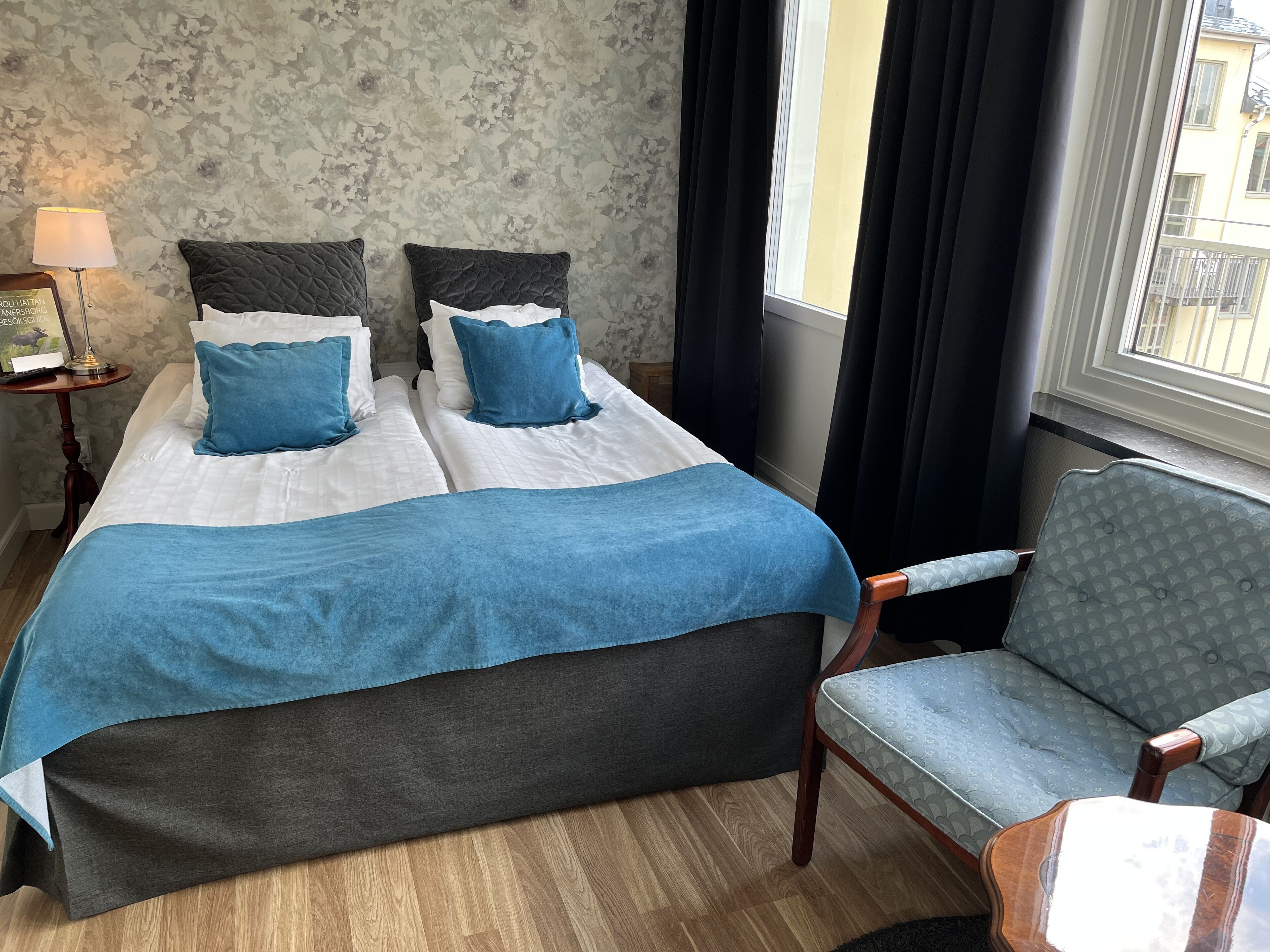 A double bed with a blue and white bedspread, two pillows, a bedside table with a lamp, and a chair with a patterned cushion.