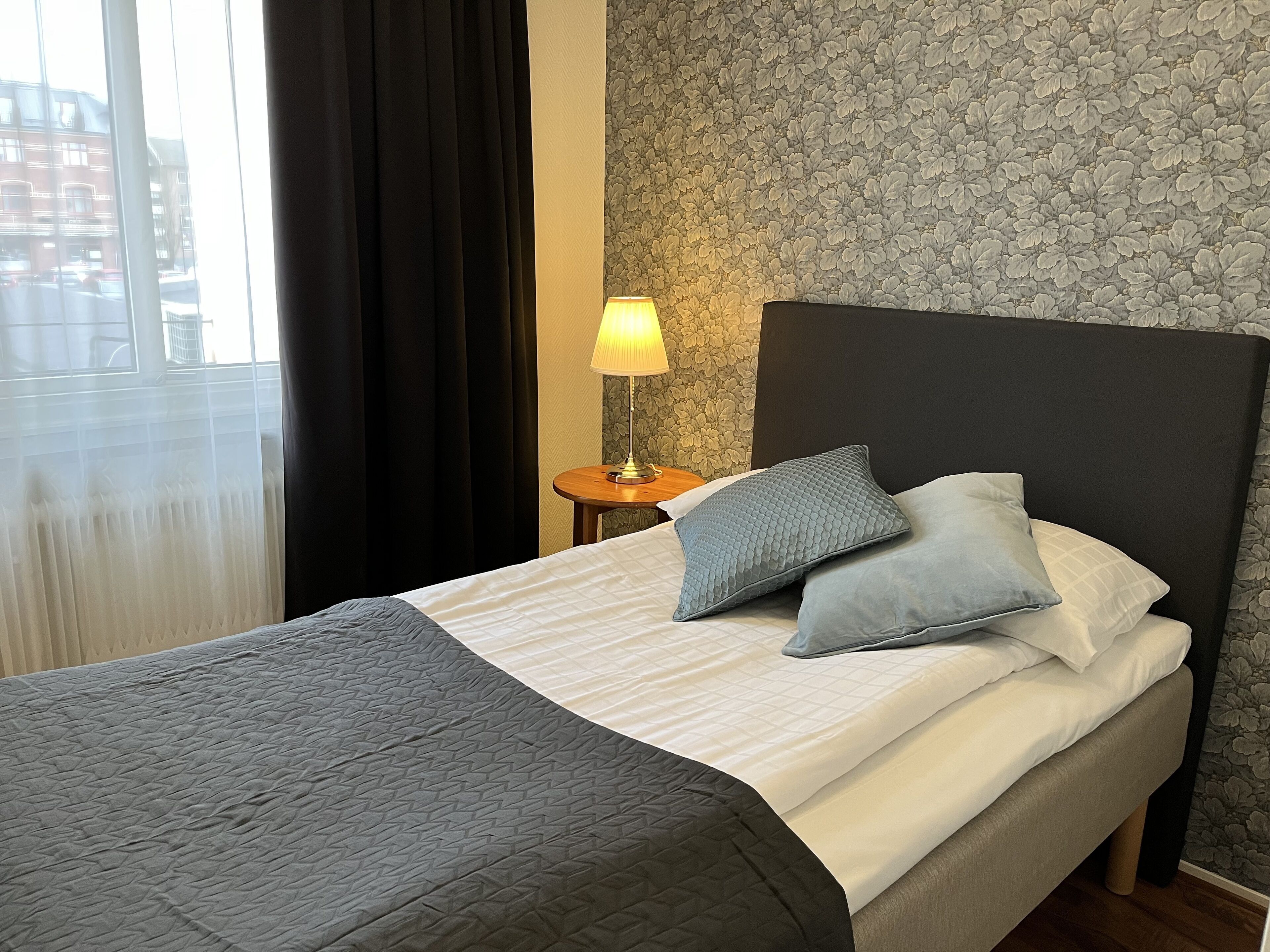 A hotel room with a bed, a nightstand with a lamp, and a window with curtains.