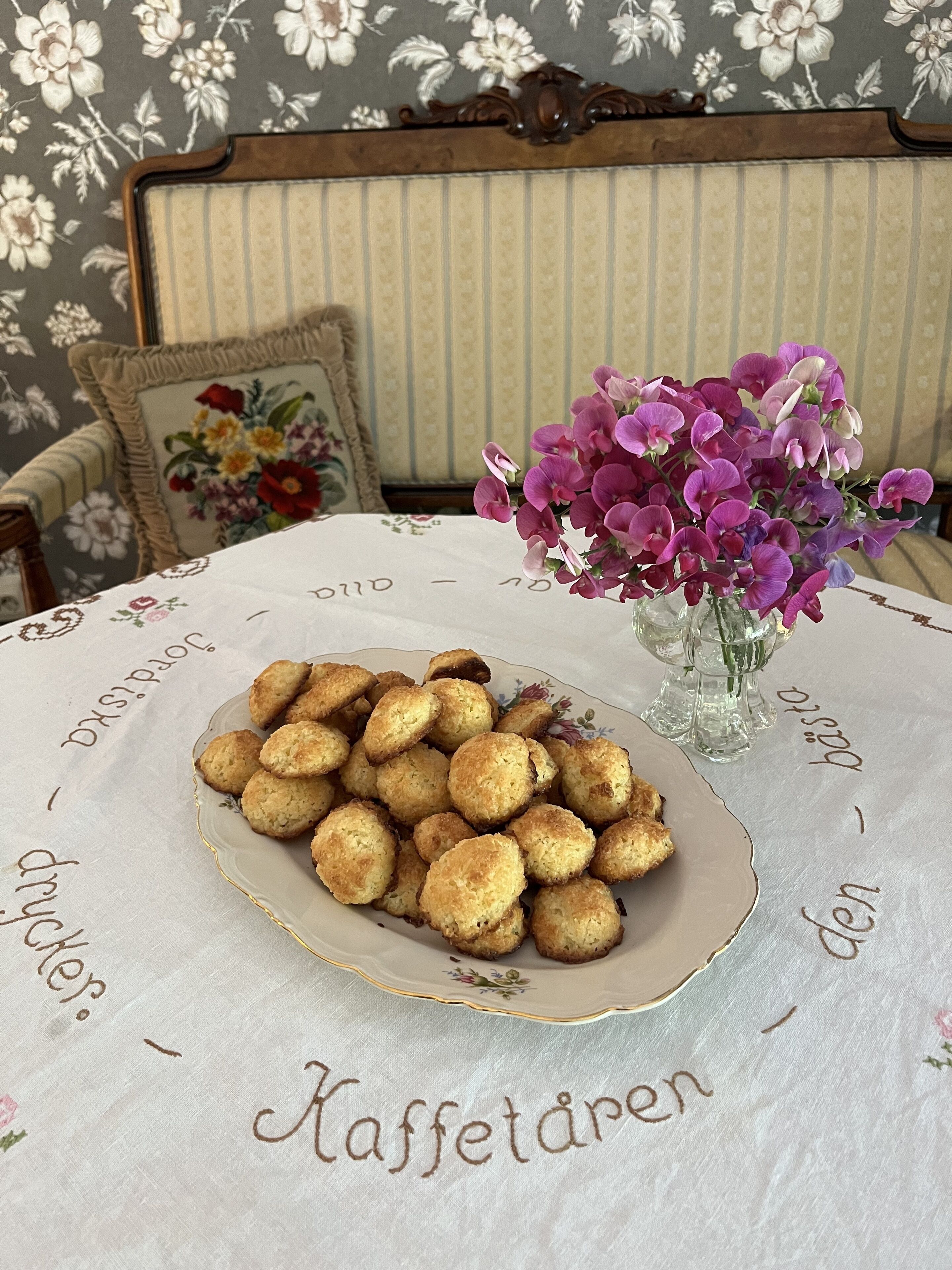 Um prato de biscoitos sobre uma mesa com toalha florida e um vaso de flores roxas.