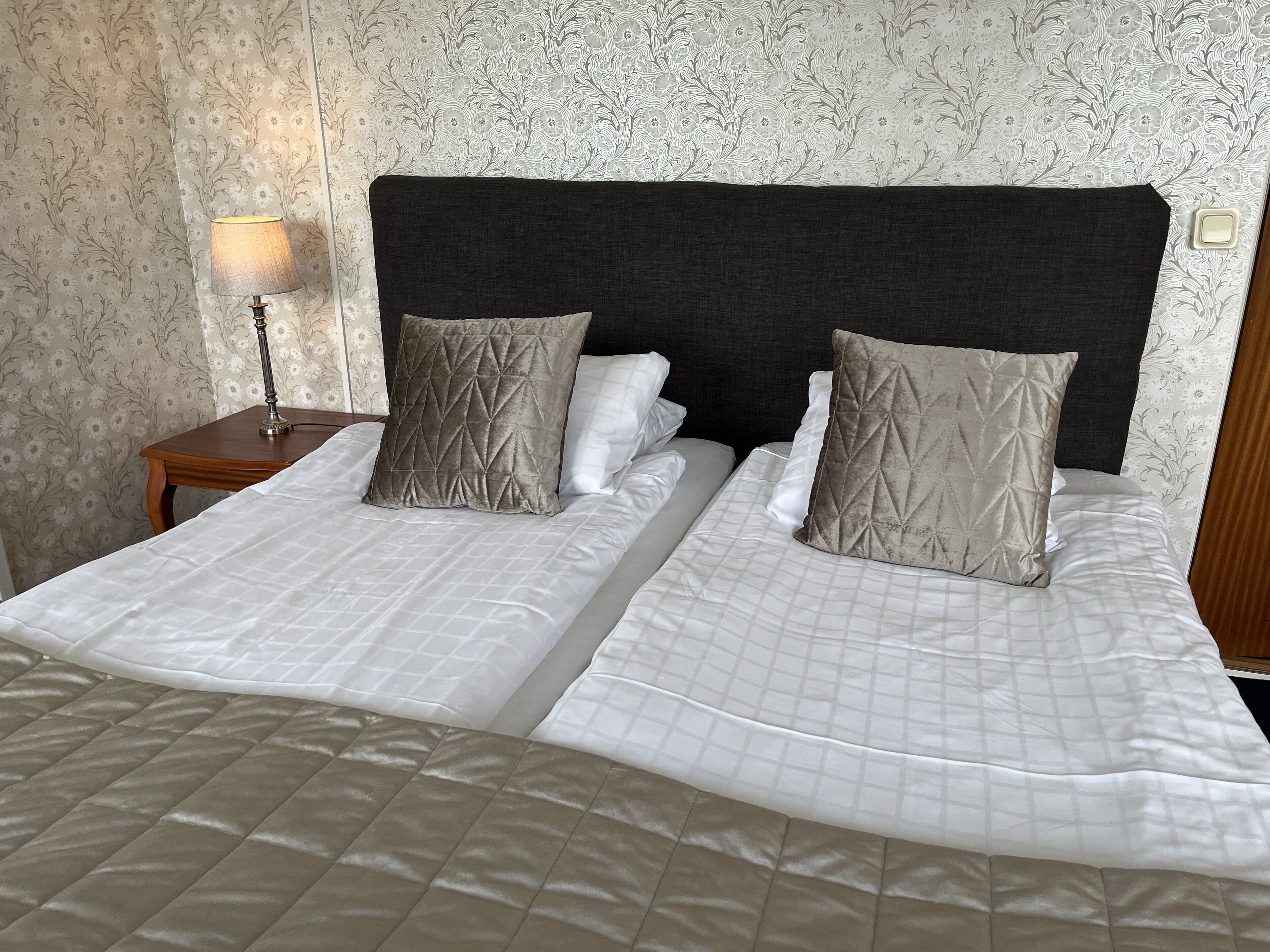 Una cama doble con cabecera acolchada, dos almohadas y una mesita de noche con lámpara.