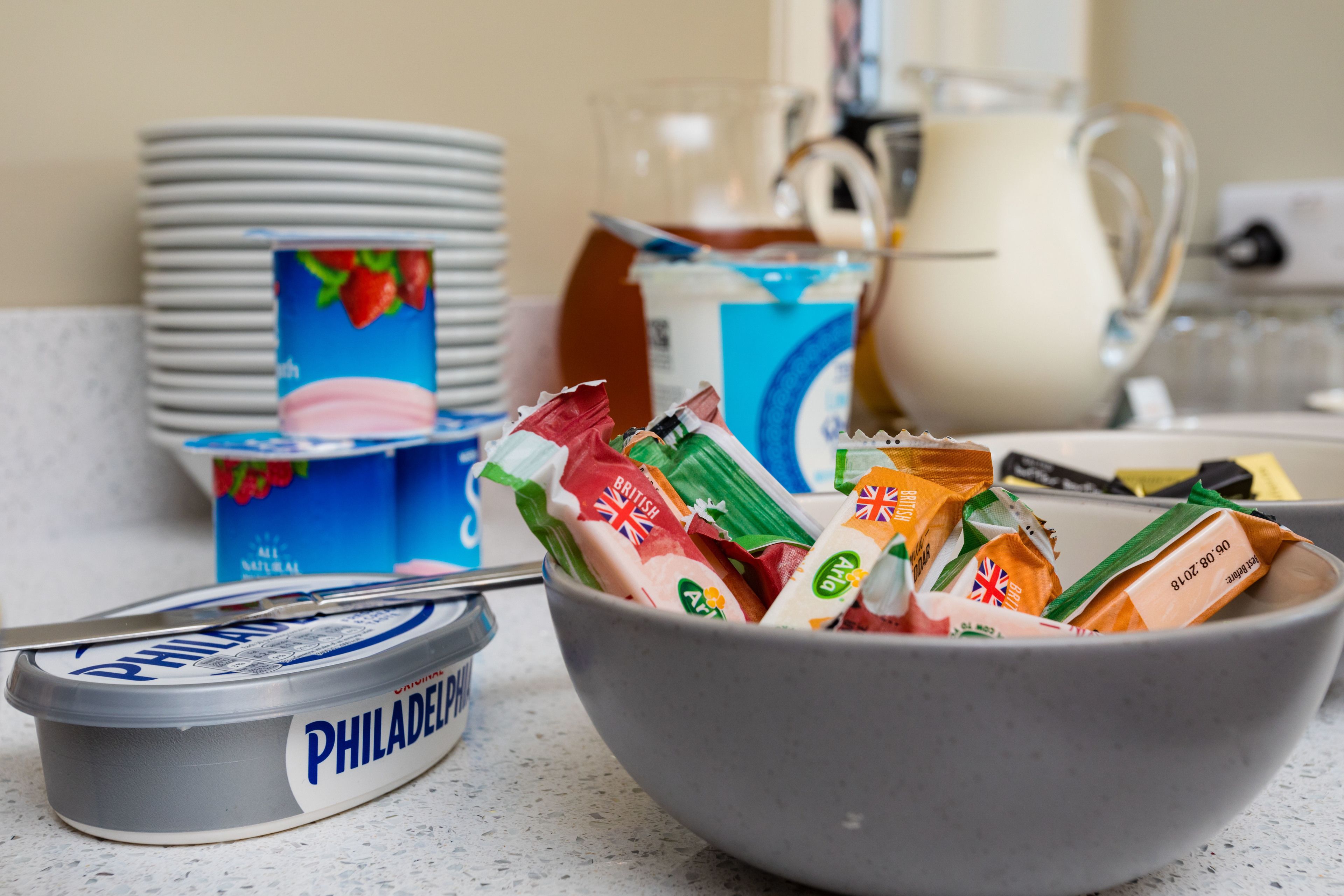 A kitchen counter with a bowl containing various packaged snacks, a container of Philadelphia ice cream, and stacks of white plates.