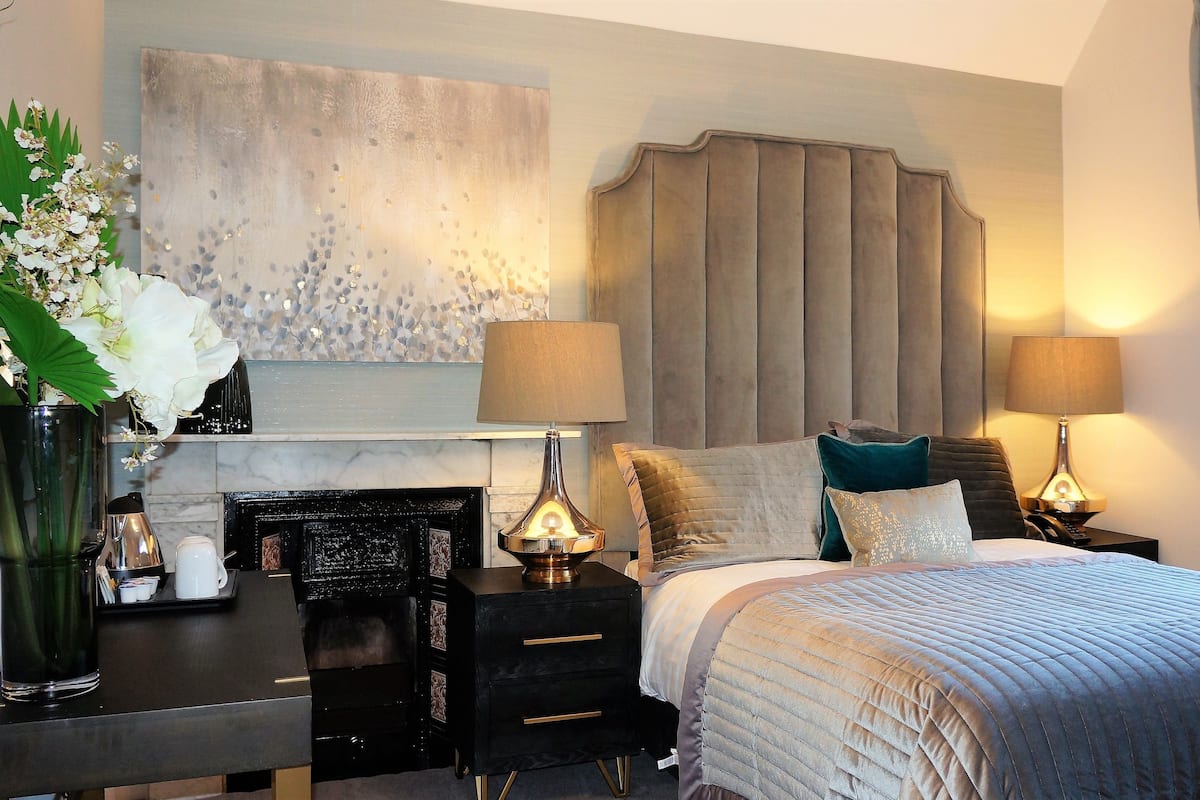 A modern hotel room with a large bed, two bedside tables with lamps, a fireplace, and a decorative wall piece.