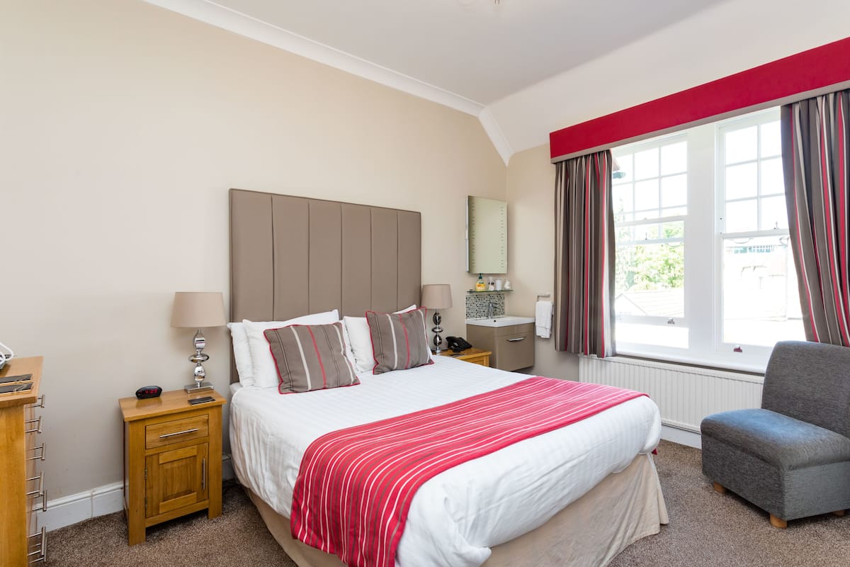 A hotel room with a large bed, bedside tables, a chair, a window with curtains, a mirror, and a small table with a lamp.
