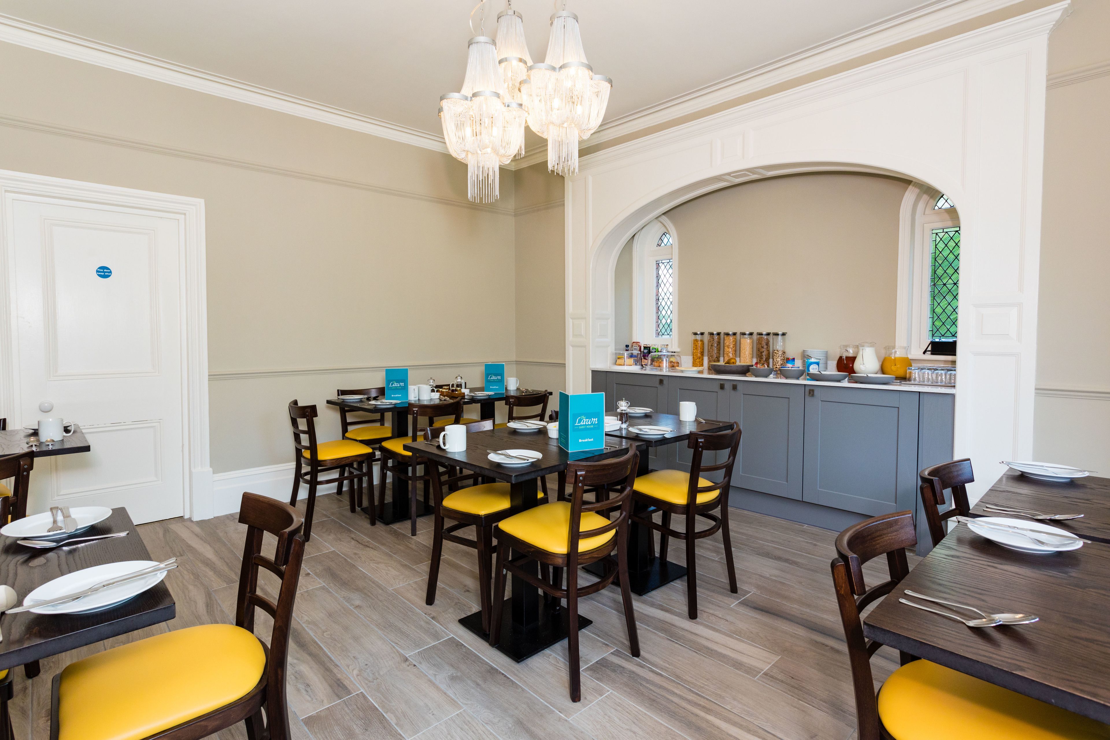 A dining area with multiple tables and chairs, a kitchen in the background, and a chandelier.