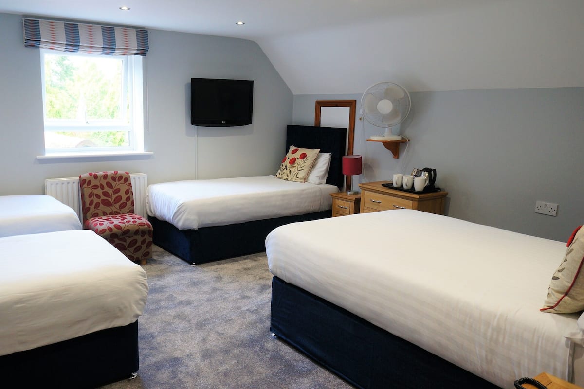 A hotel room with three single beds, a chair, a TV, a fan, and a window with curtains.
