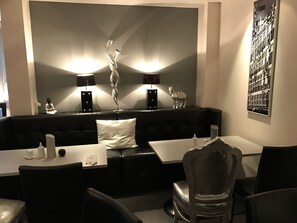 A modern dining area with a black leather booth, white tables, and black chairs. There are two wall-mounted lamps, a decorative sculpture, and a framed picture on the wall. - Hotel Lindenhof (Düsseldorf)