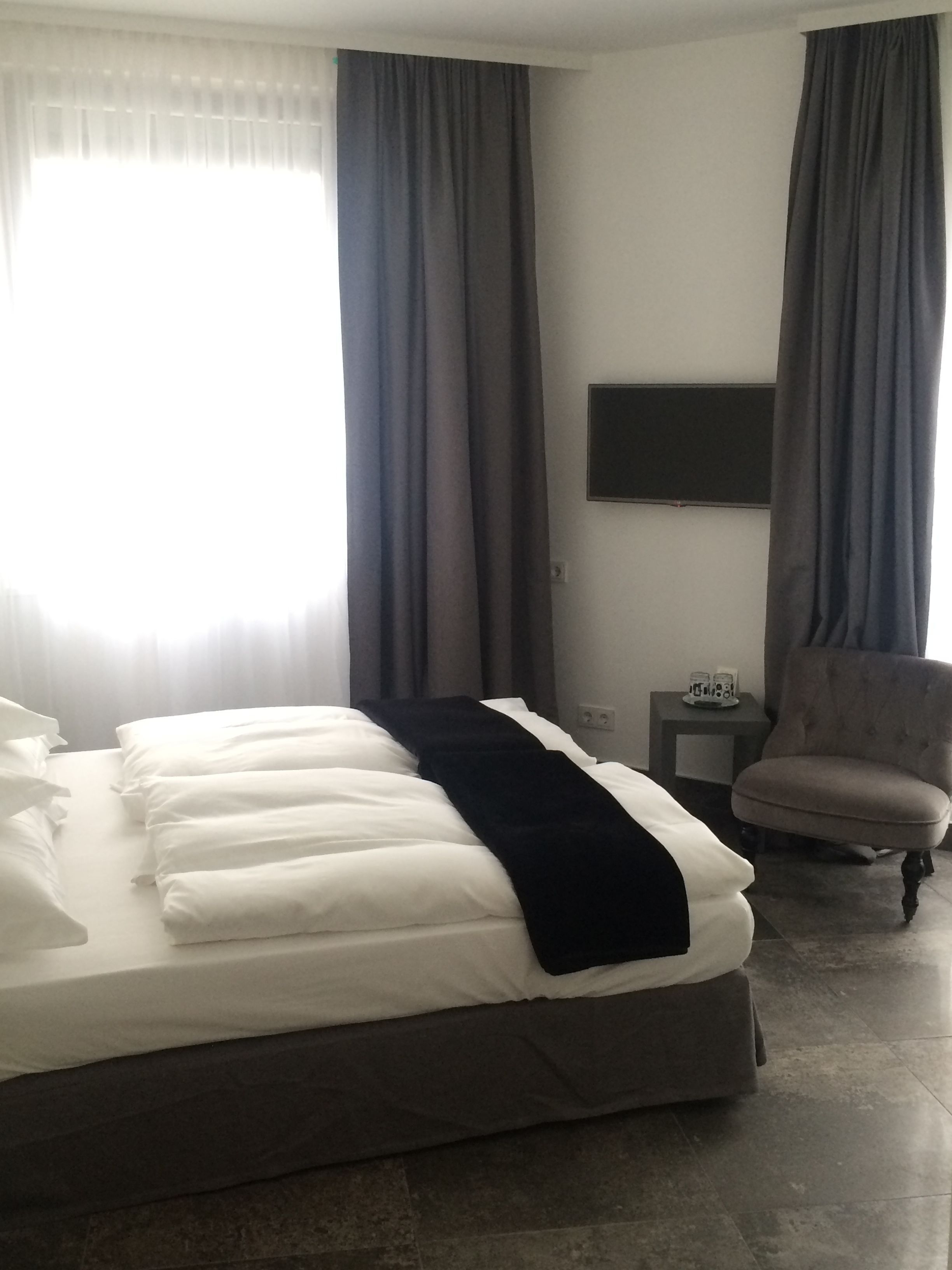 A hotel room with a bed, a chair, a small table, and a flat-screen TV mounted on the wall.