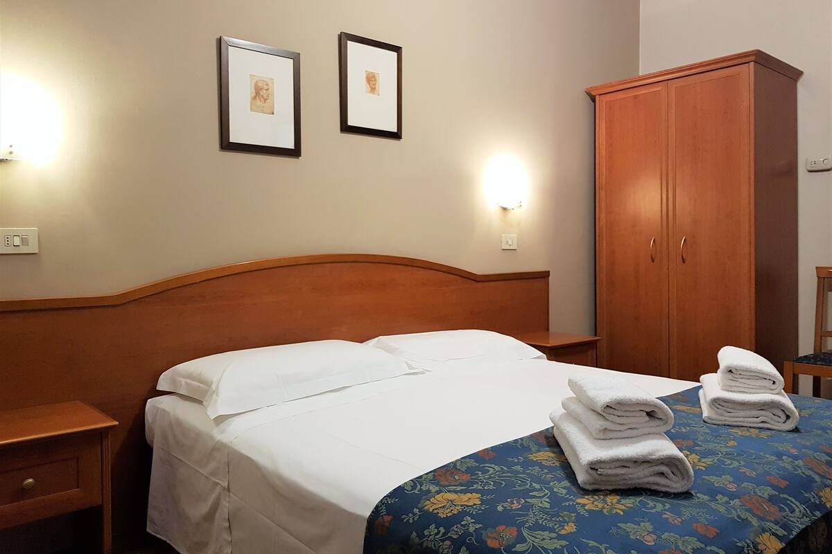 Une chambre d’hôtel avec un lit doté d’une tête de lit en bois, du linge de lit blanc, une couvre-lit à motifs floraux bleus, deux tableaux encadrés au mur et une armoire en bois.