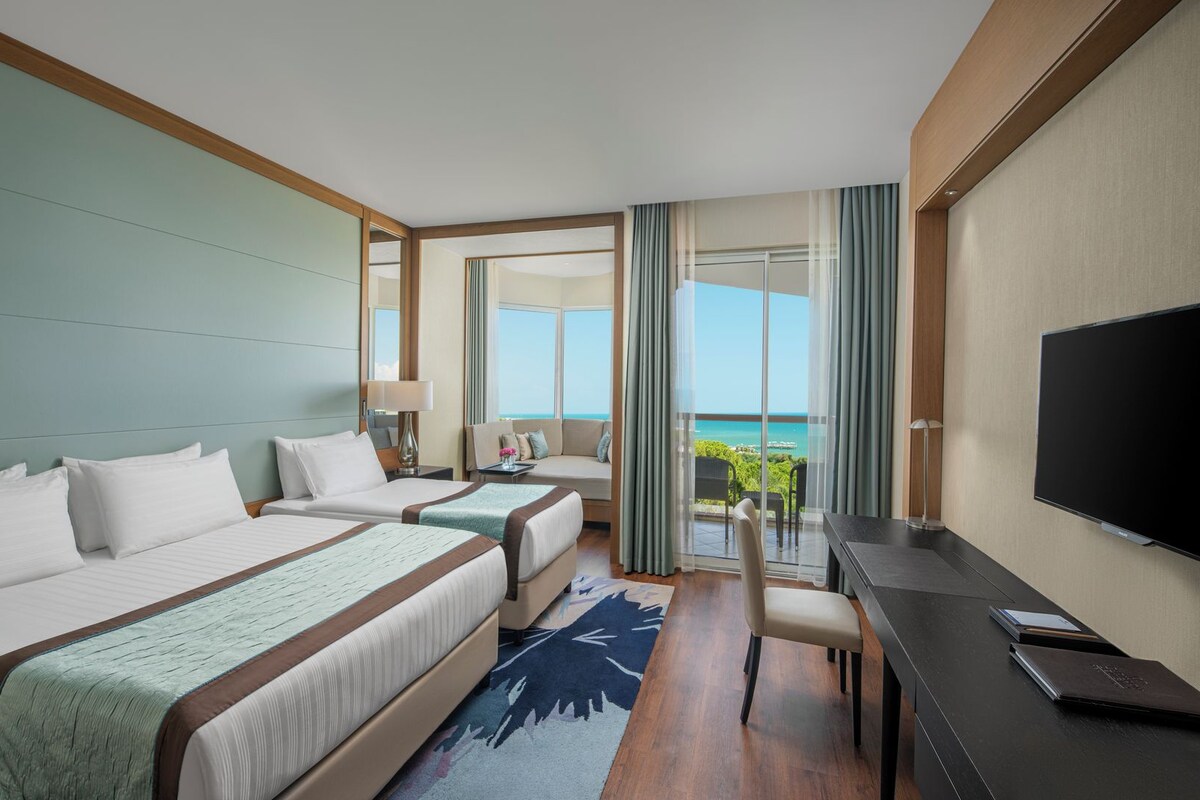 A hotel room with two beds, a desk, a TV, and a balcony with a view of the sea.