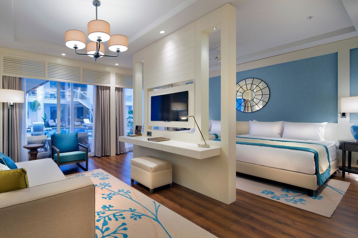 A modern hotel room with a sofa, a desk, a bed, a TV, and a large window with curtains.