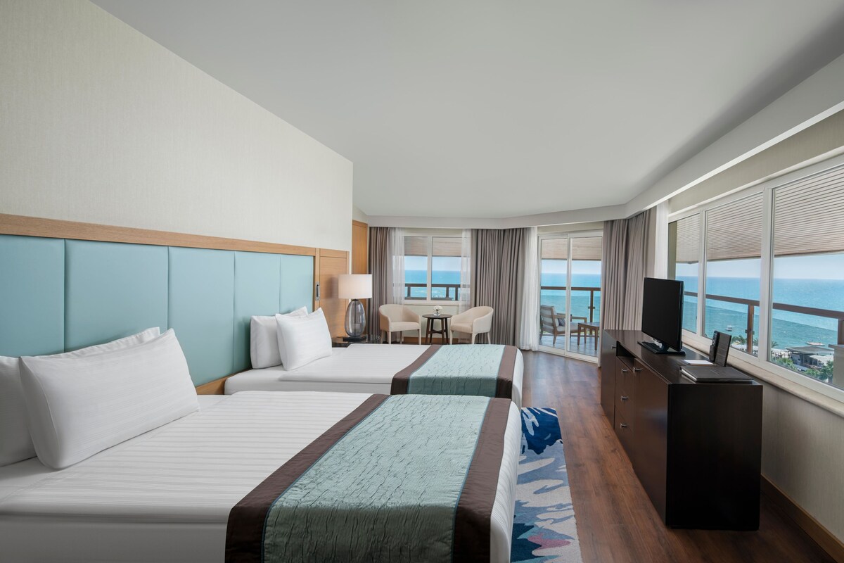 A hotel room with two beds, a desk, a TV, and a balcony with a view of the sea.