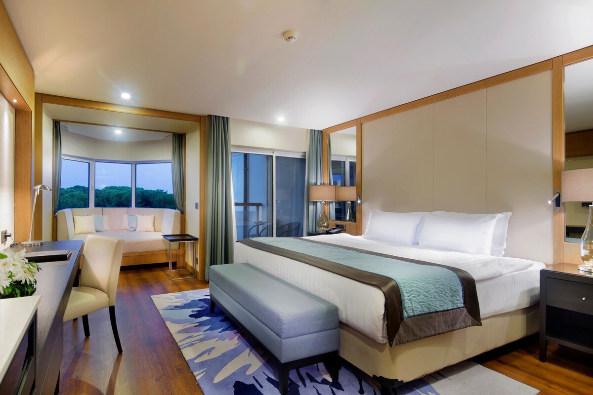 A modern hotel room with a large bed, a desk with a chair, a small table, and a large window offering a view of the outdoors.