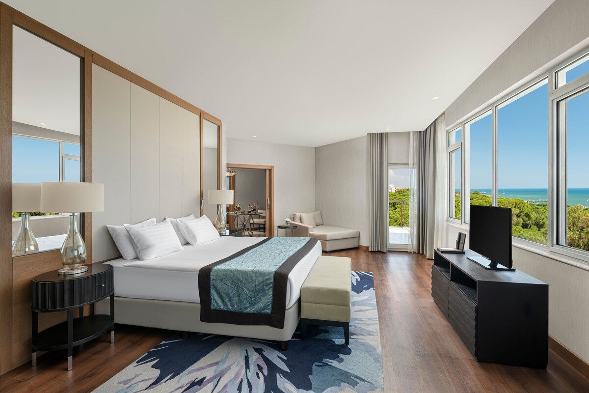 A modern hotel room with a large bed, a seating area, a TV, and a view of the ocean.