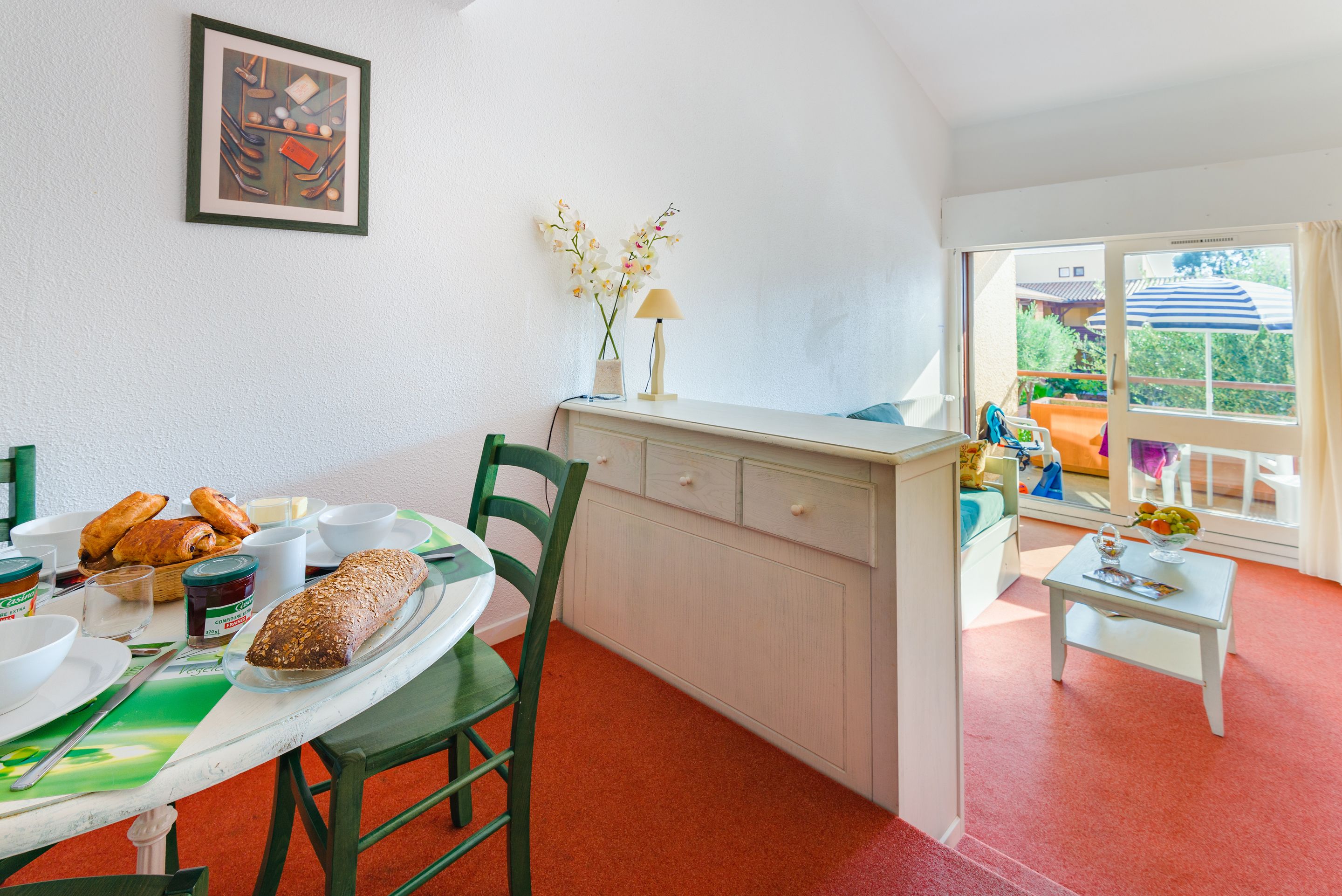 A dining area with a table set for breakfast, a green chair, a white cabinet, and a window with a view of the outdoors.