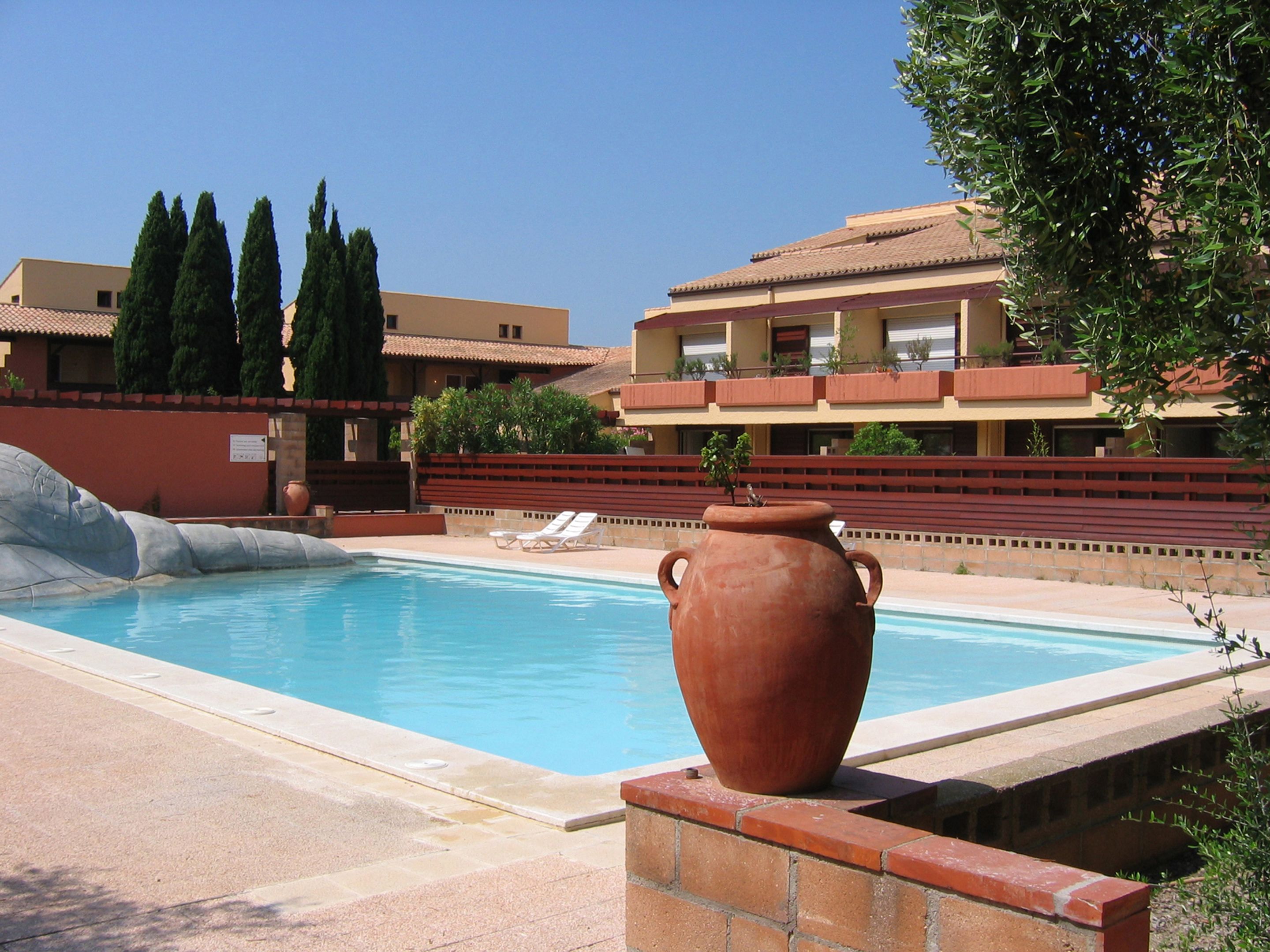 A swimming pool with a large terracotta pot, a red wall, and a building with balconies and trees in the background.