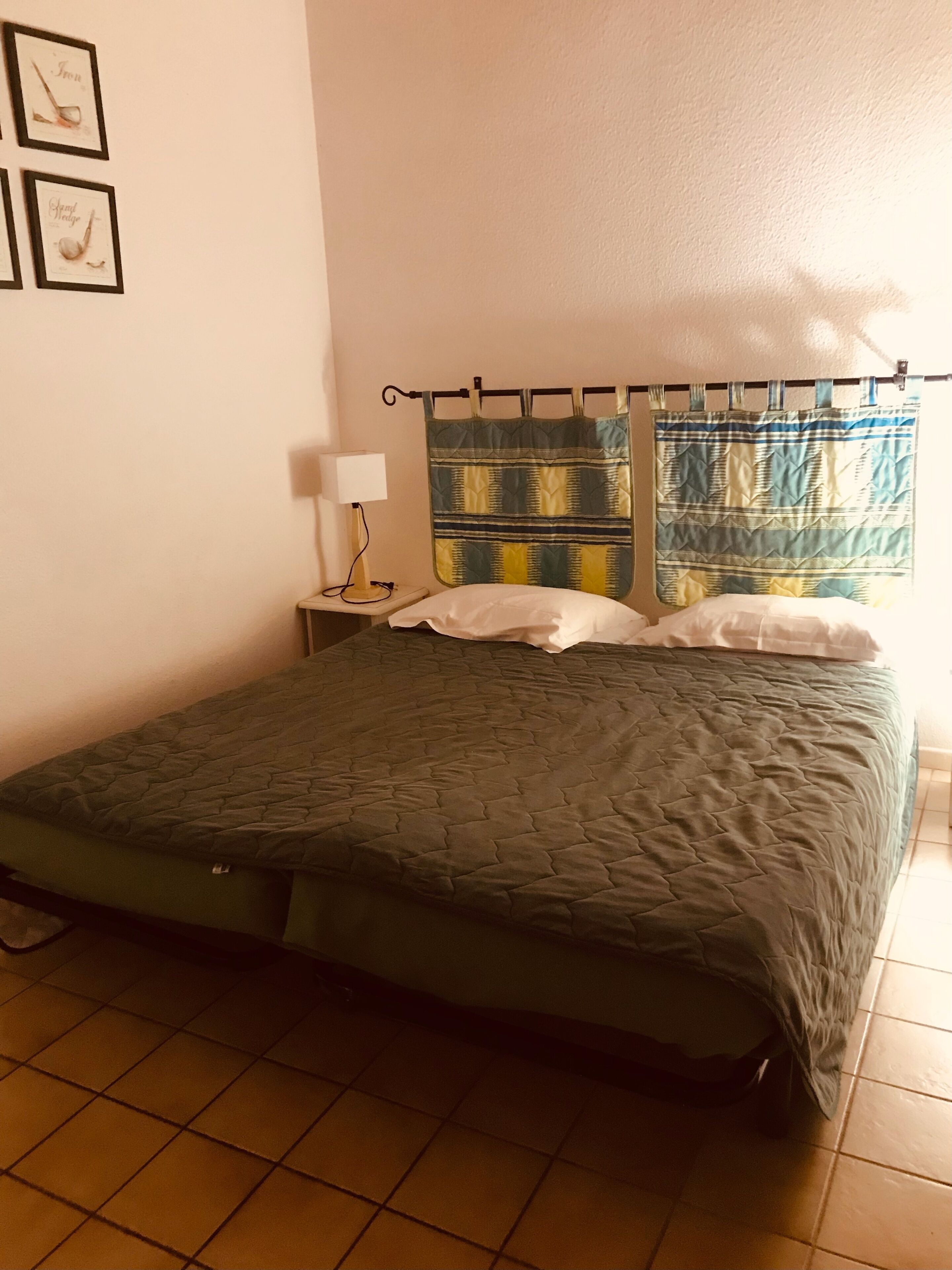 A single bed with a quilt, a nightstand with a lamp, a window with curtains, and framed pictures on the wall.