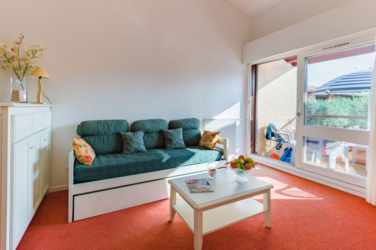Ein Zimmer mit einem türkisblauen Sofa, einem weißen Couchtisch mit Obstschale, einem weißen Schrank und einem großen Fenster mit Blick auf Grünflächen und ein Gebäude.