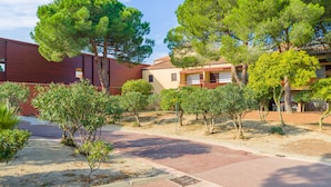A courtyard with a paved walkway, surrounded by trees and a building with a red exterior. - Résidence Lagrange Vacances du Golf (Saint-Cyprien)
